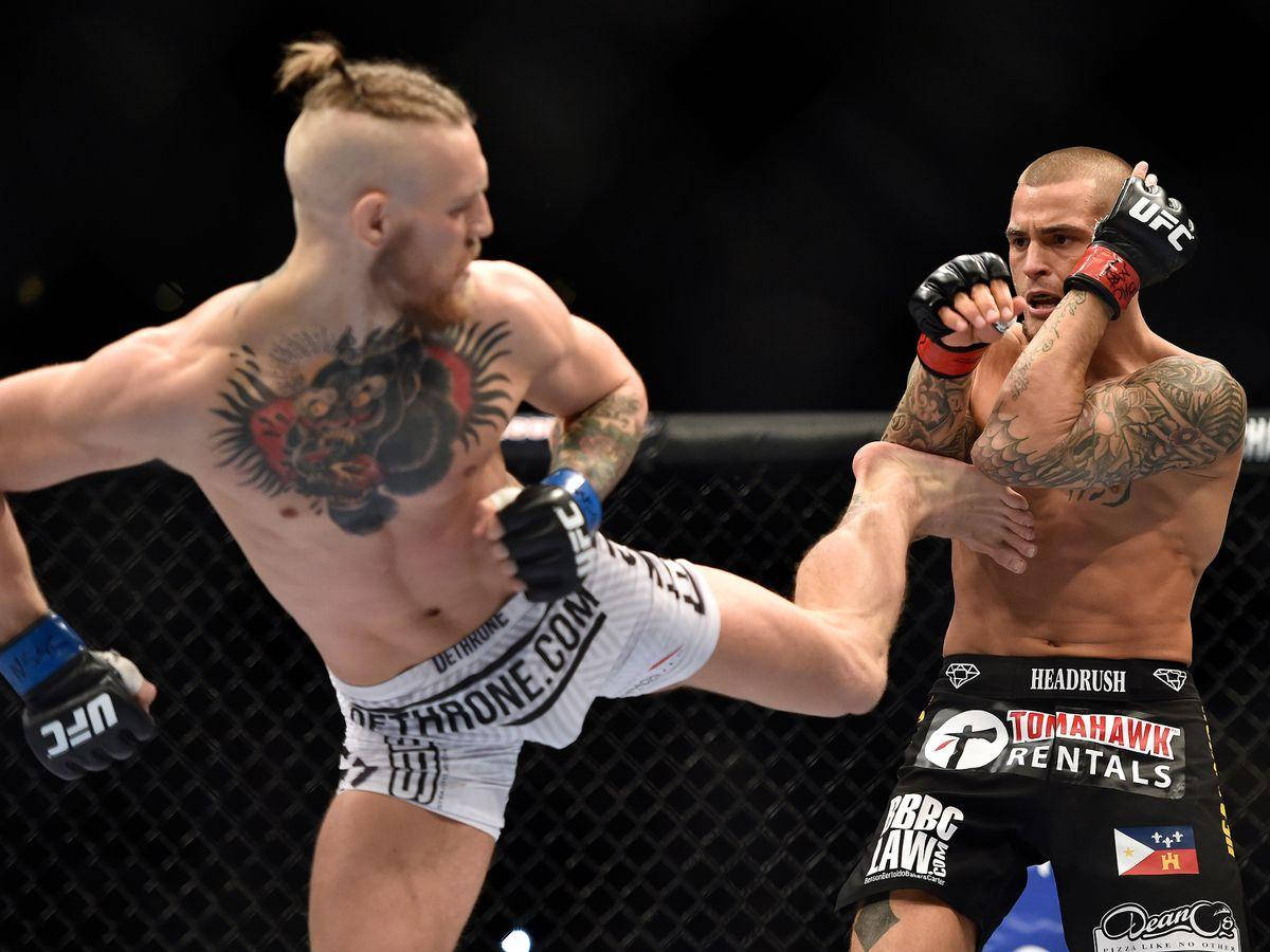 UFC 桌布 壁紙 — 兩名有刺青的MMA拳手在八角籠中交鋒，其中一人踢出高踢，另一人在明亮的場館燈光下阻擋。