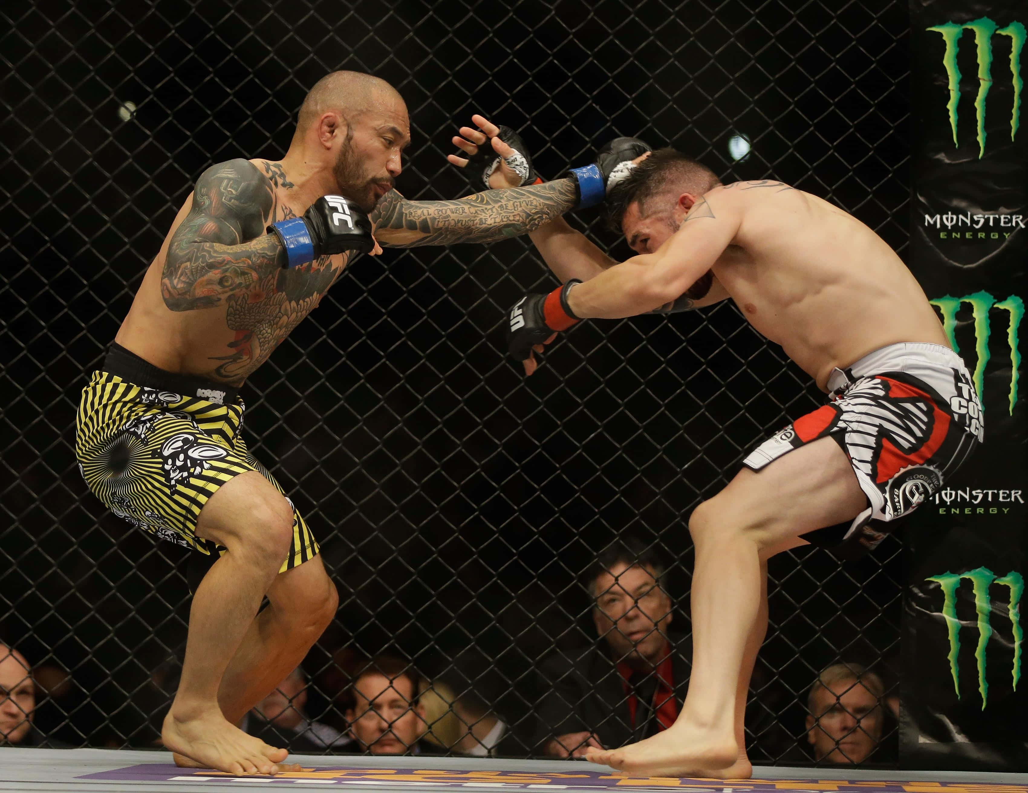 UFC 桌布 壁紙 — 兩名有刺青的MMA拳手在籠中交鋒，一人出拳，另一人在昏暗的場地中阻擋。