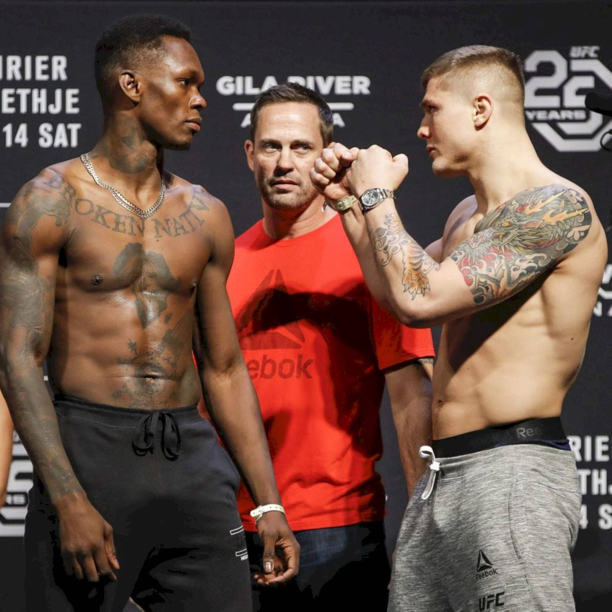 UFC 桌布 壁紙 — 兩名有刺青的UFC拳手在明亮的場館燈光下，在過磅時對峙，中間有裁判。
