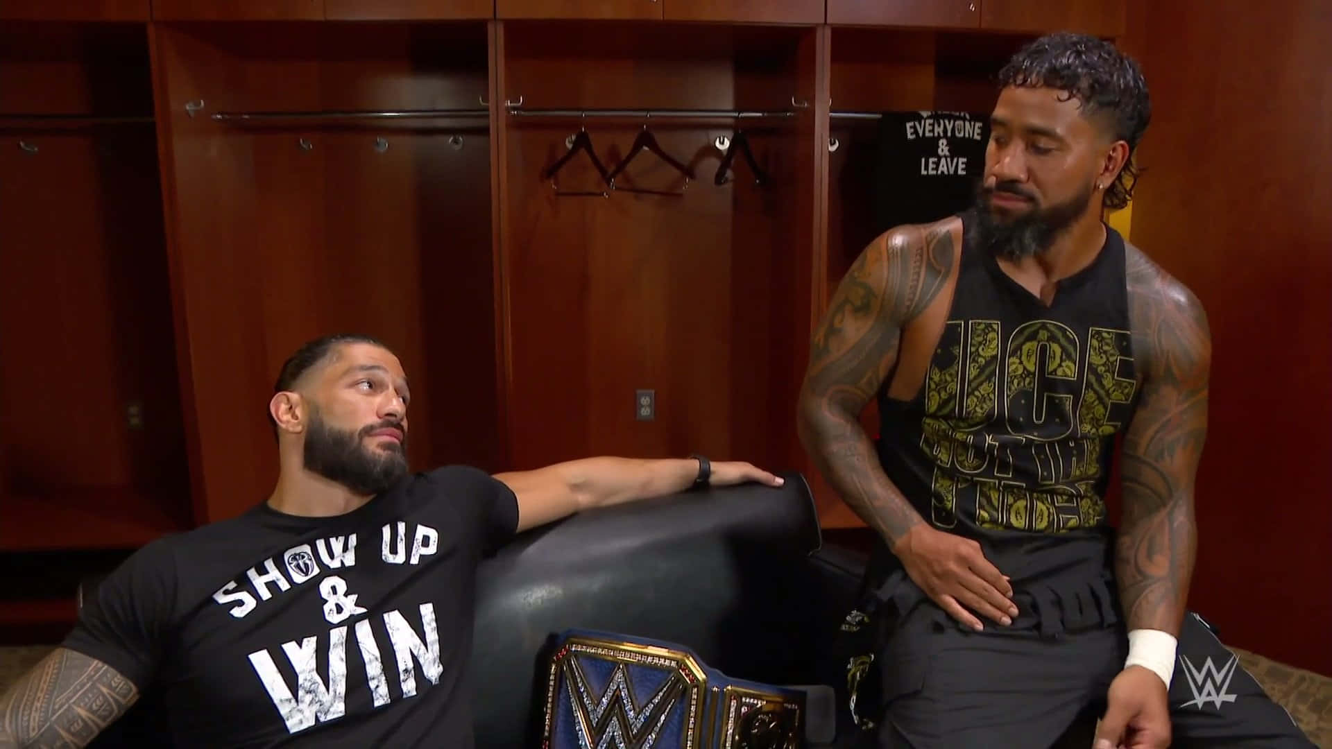 Roman Reigns 桌布 壁紙 — 兩名有刺青的WWE摔角手坐在木製更衣室裡，其中一人靠在冠軍腰帶旁邊的沙發上。