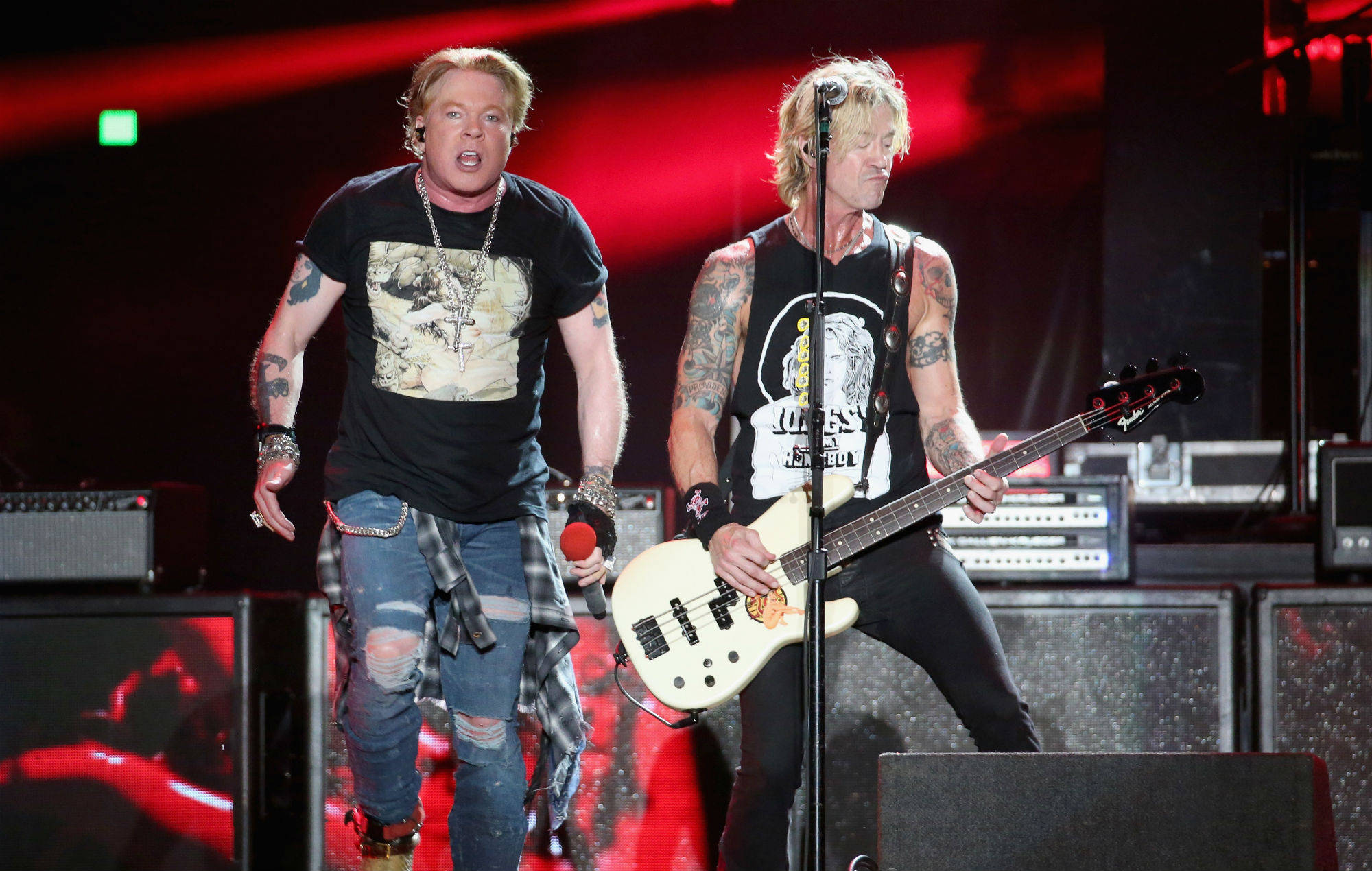 guns n roses 桌布 壁紙 — 兩名有刺青的搖滾音樂家在紅燈下，帶著擴音器和低音吉他，在舞台上充滿活力地表演。