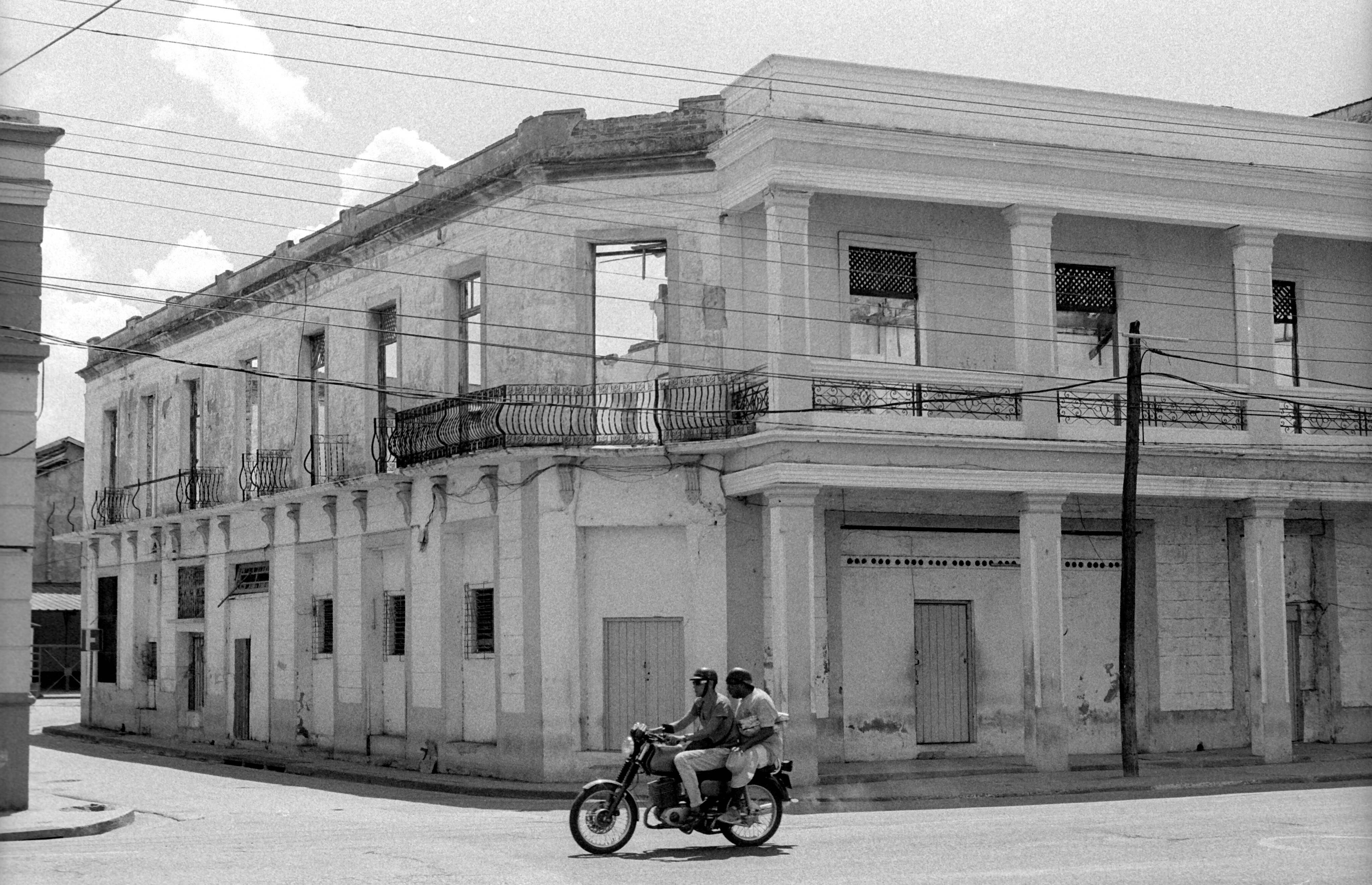 Cuba 桌布 壁紙 — 兩名機車騎士在陰天下駛過一棟破舊的殖民時期建築，建築物窗戶破損，陽台華麗。