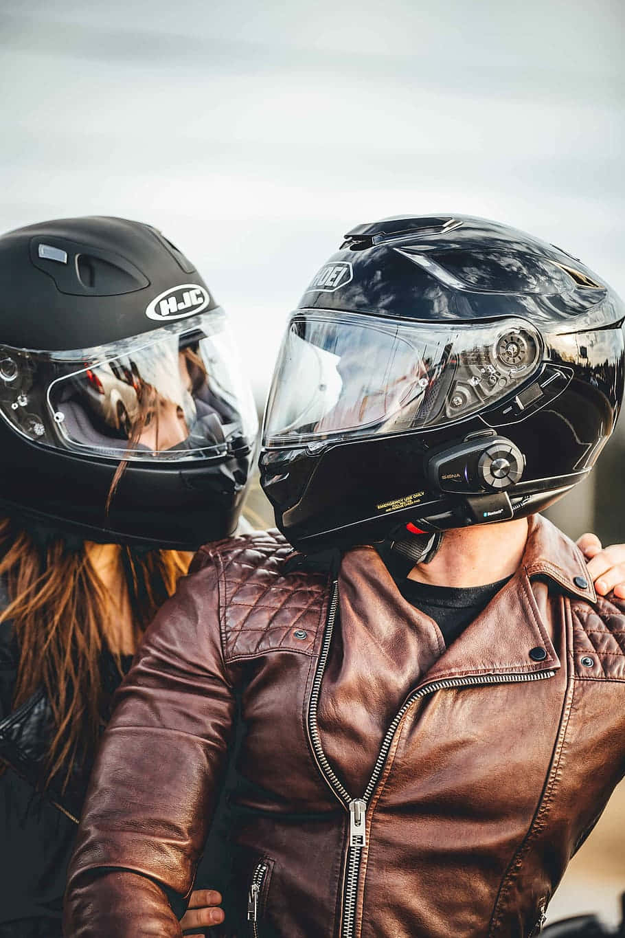 Lovers 桌布 壁紙 — 兩名機車騎士戴著黑色安全帽和棕色皮夾克，在柔和的灰色天空下擁抱。
