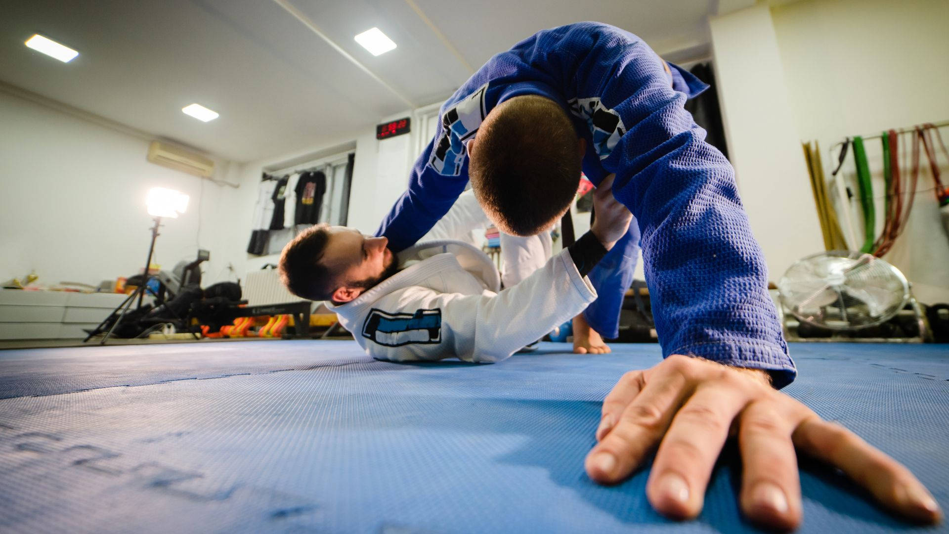 martial arts 桌布 壁紙 — 兩名武術家在光線充足的健身房的藍色軟墊上激烈纏鬥，背景有訓練器材。