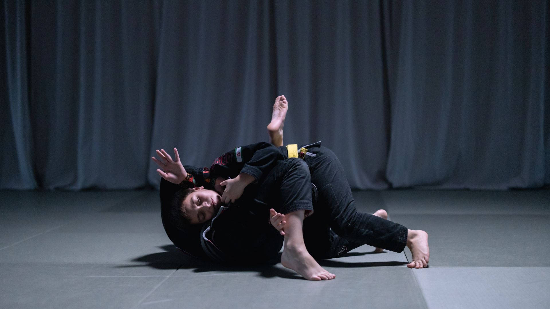 martial arts 桌布 壁紙 — 兩名武術家在灰色軟墊上激烈纏鬥，背景為深色窗簾，燈光效果戲劇化。