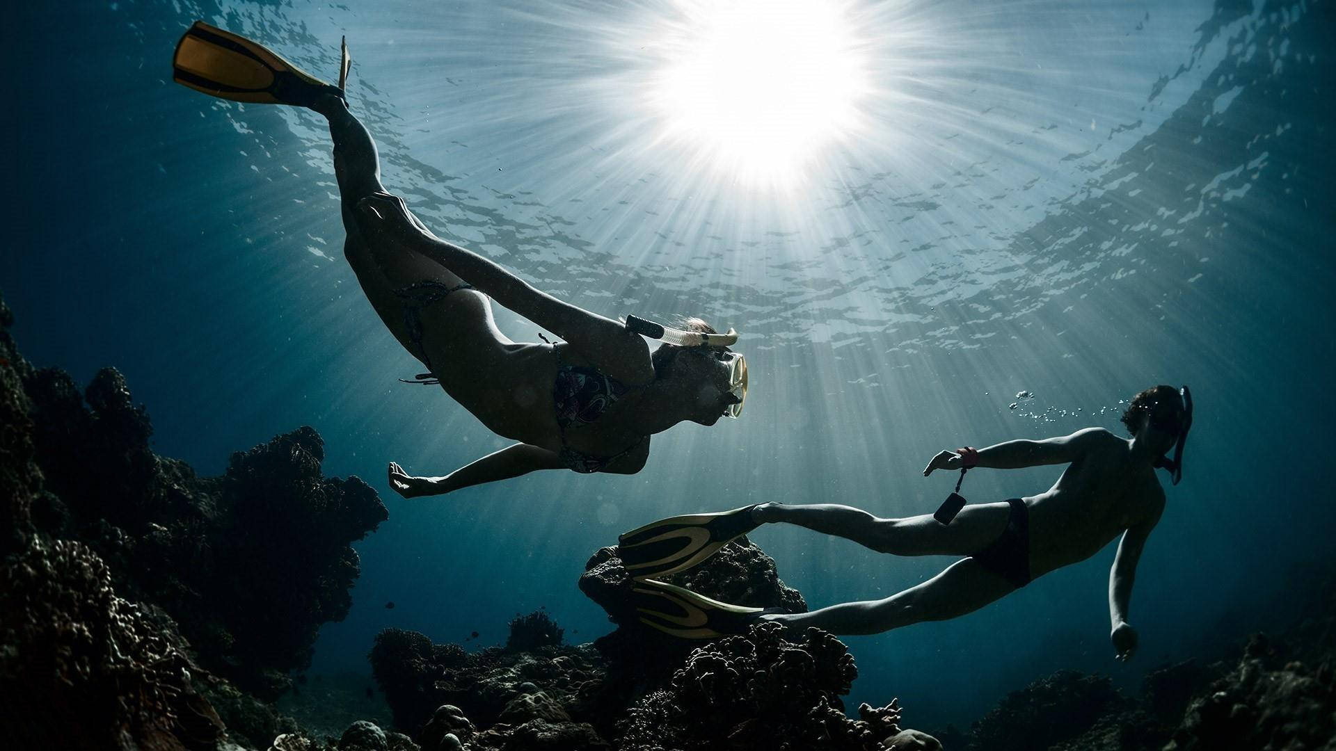 Snorkeling 桌布 壁紙 — 兩名浮潛者在水下優雅地滑行，陽光穿過海面照射下來，照亮了下方的珊瑚礁。