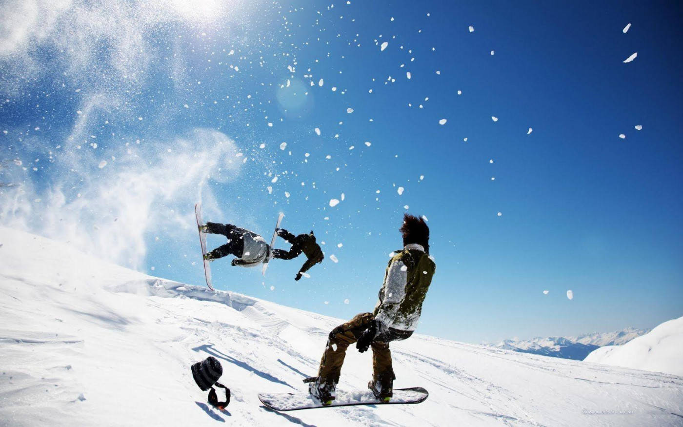 雪 桌布 壁紙 — 兩名滑雪板運動員在半空中，背景是明亮的藍天，雪花飛濺，一人落地，另一人在雪坡上觀看。