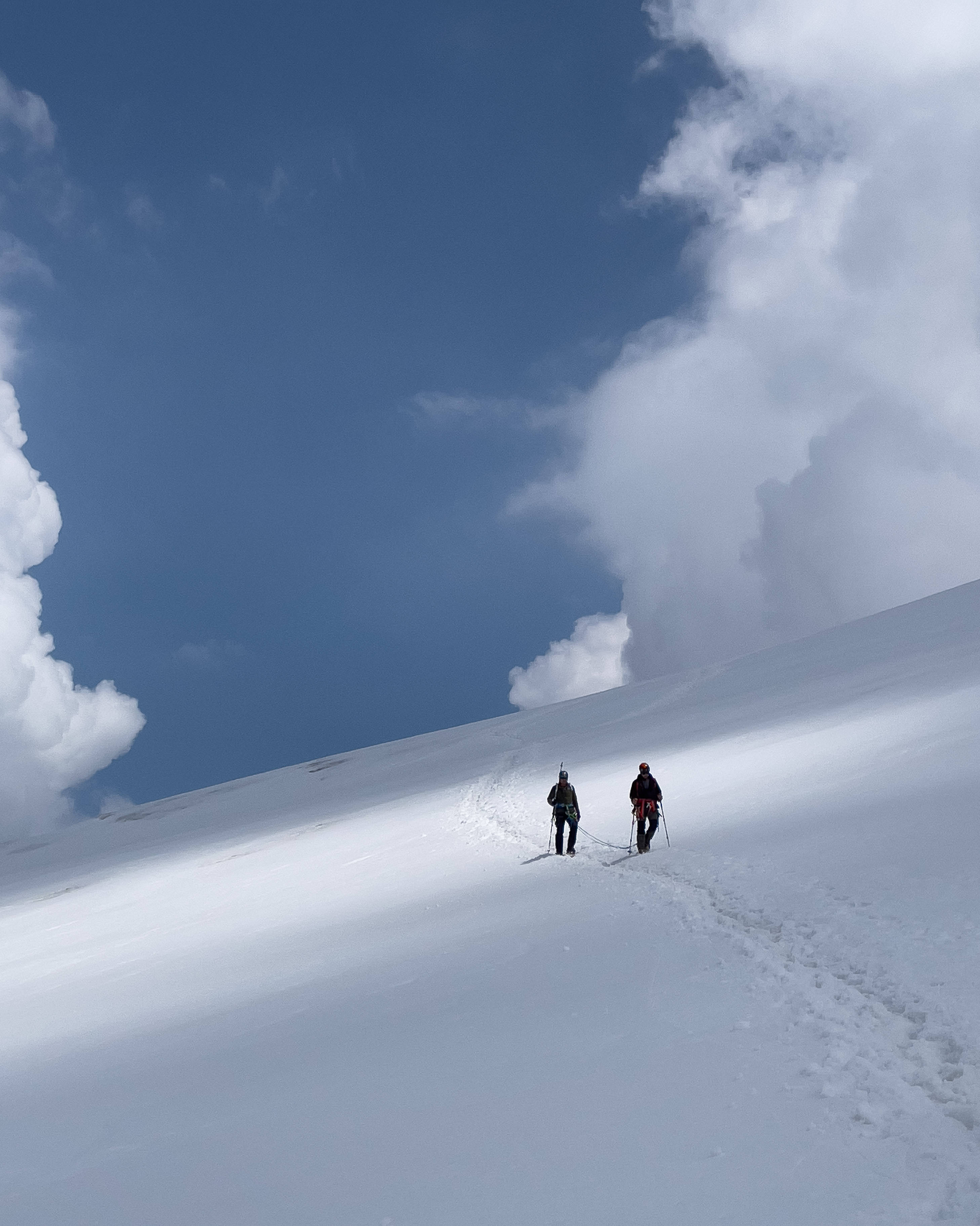 最佳桌面 桌布 壁紙 — 兩名滑雪者在一片廣闊的雪山斜坡上攀登，天空是戲劇性的藍色，點綴著蓬鬆的白雲。