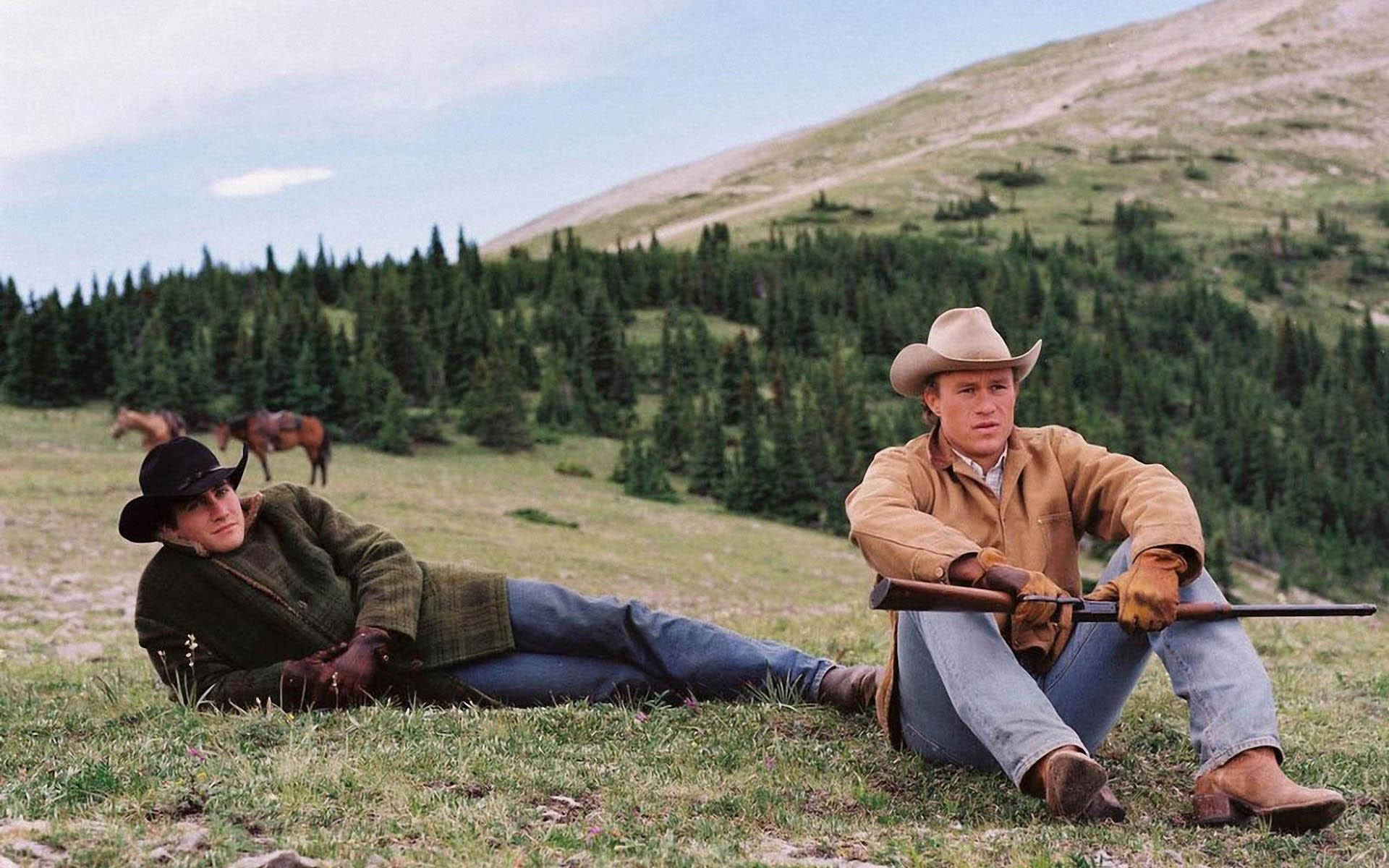 brokeback mountain 桌布 壁紙 — 兩名牛仔在長滿草的山坡上放鬆，馬匹在他們身後於部分多雲的天空下吃草。