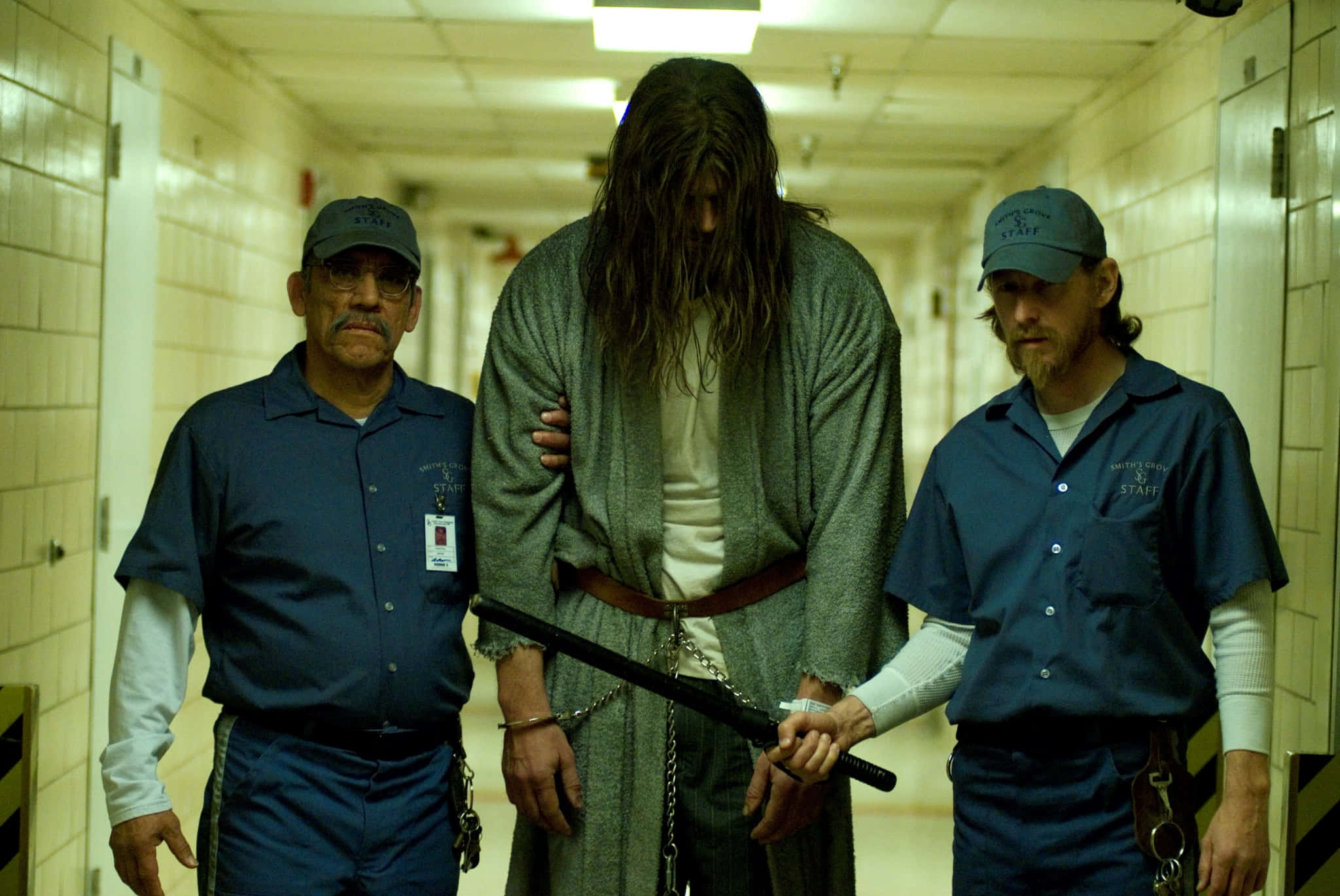 Prison 桌布 壁紙 — 兩名獄警護送一名身穿灰色長袍的長髮囚犯，沿著螢光燈照亮的機構走廊前行。