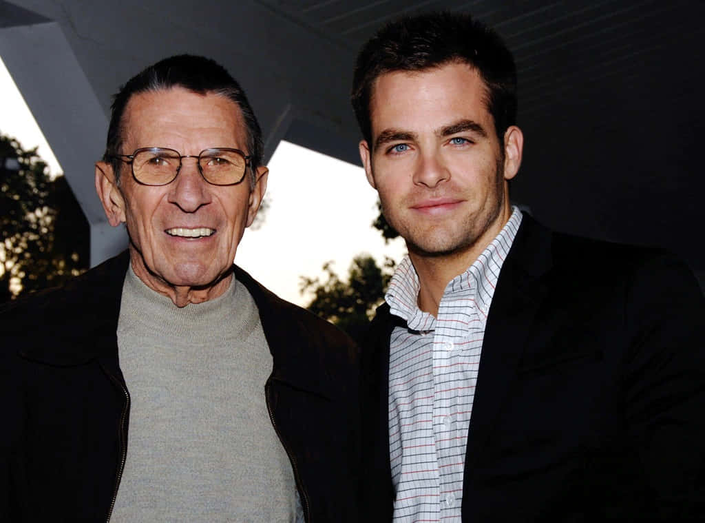 chris pine 桌布 壁紙 — 兩名男子，一位戴眼鏡較年長，另一位藍眼睛較年輕，在現代建築結構下並肩微笑站立。