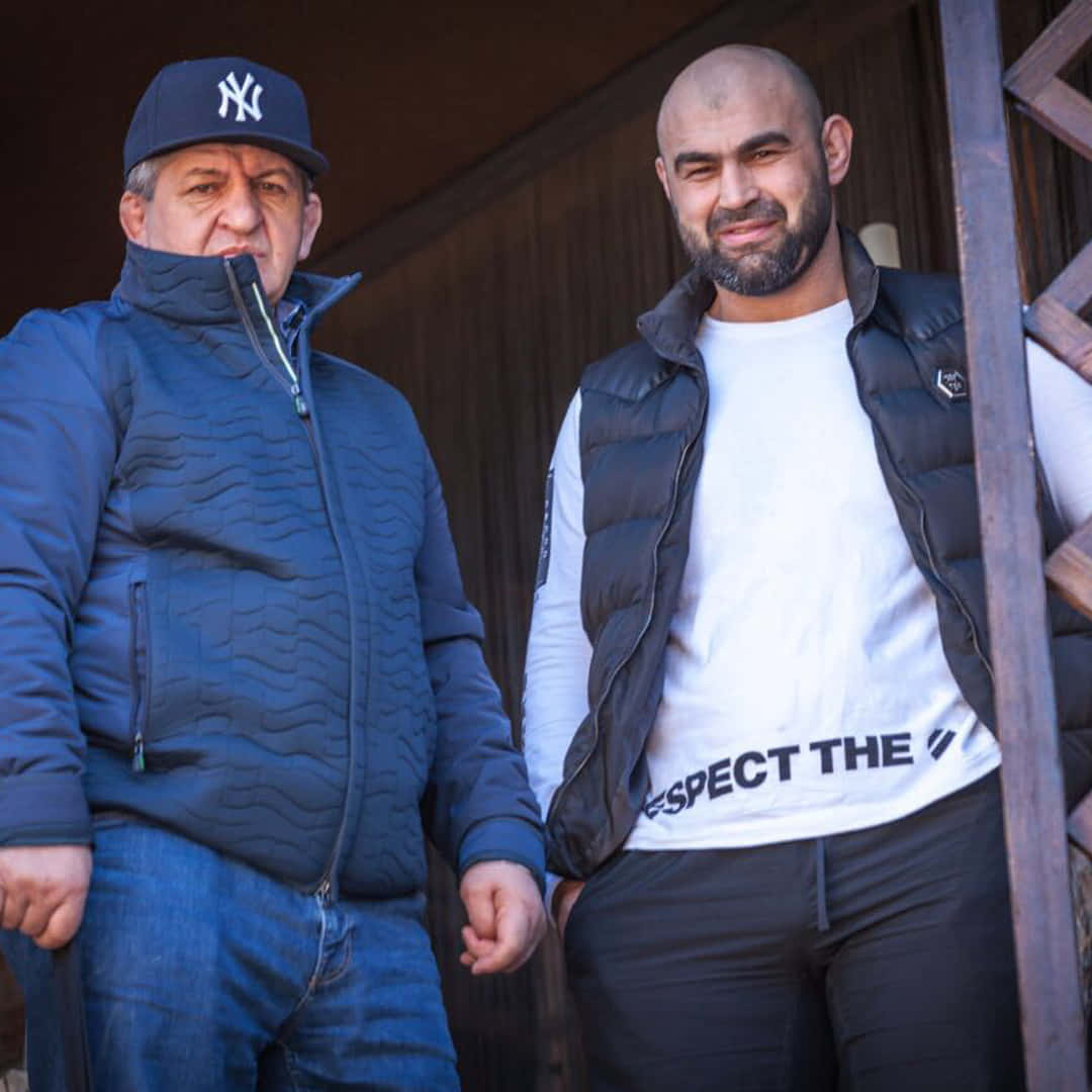 MMA 桌布 壁紙 — 兩名男子並排站在木結構前，一人穿著藍色絎縫外套和洋基隊帽，另一人穿著黑色背心，內搭白色T恤，上面有文字。