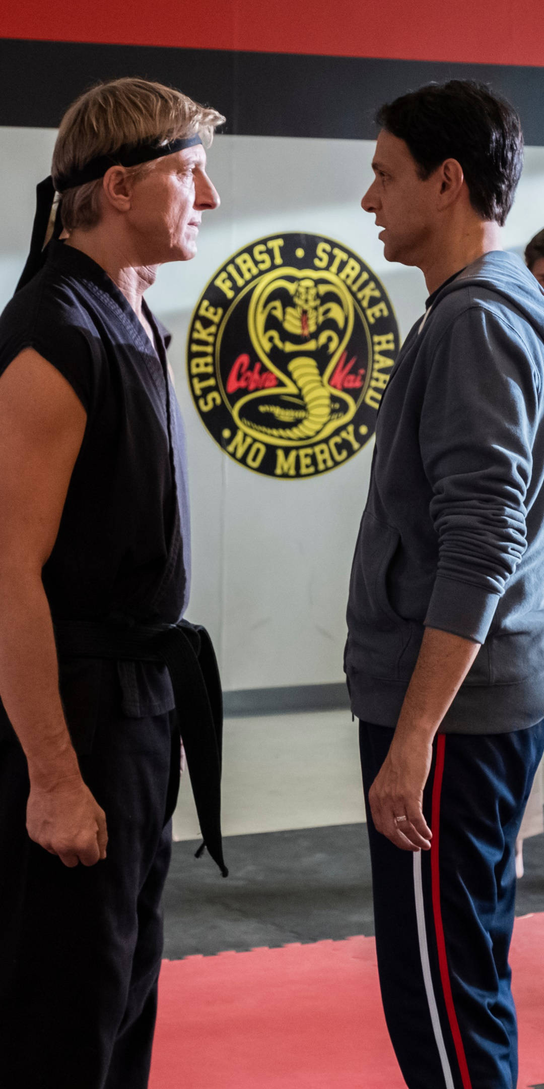 眼鏡蛇道館 桌布 壁紙 — 兩名男子在一個有 Cobra Kai 標誌、紅色軟墊和黑色牆壁的武術道場中對峙。