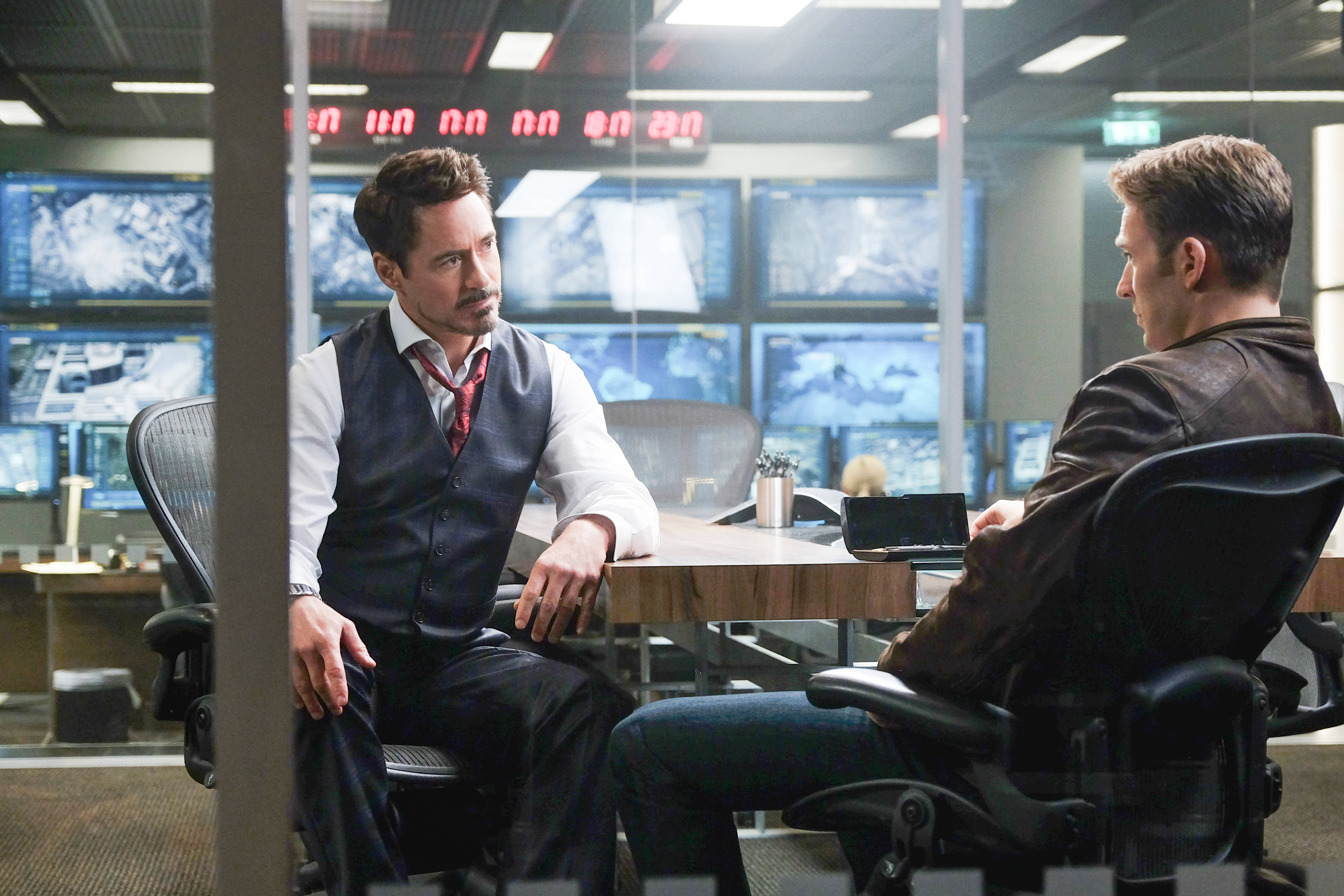 tony stark 桌布 壁紙 — 兩名男子在一個高科技指揮中心內，隔著一張被發光螢幕包圍的木桌進行嚴肅對話。