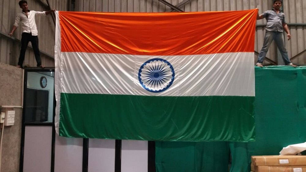 Indian 桌布 壁紙 — 兩名男子在倉庫中手持一面巨大的印度國旗，旗上有藏紅花、白色和綠色條紋以及藍色的阿育王法輪。