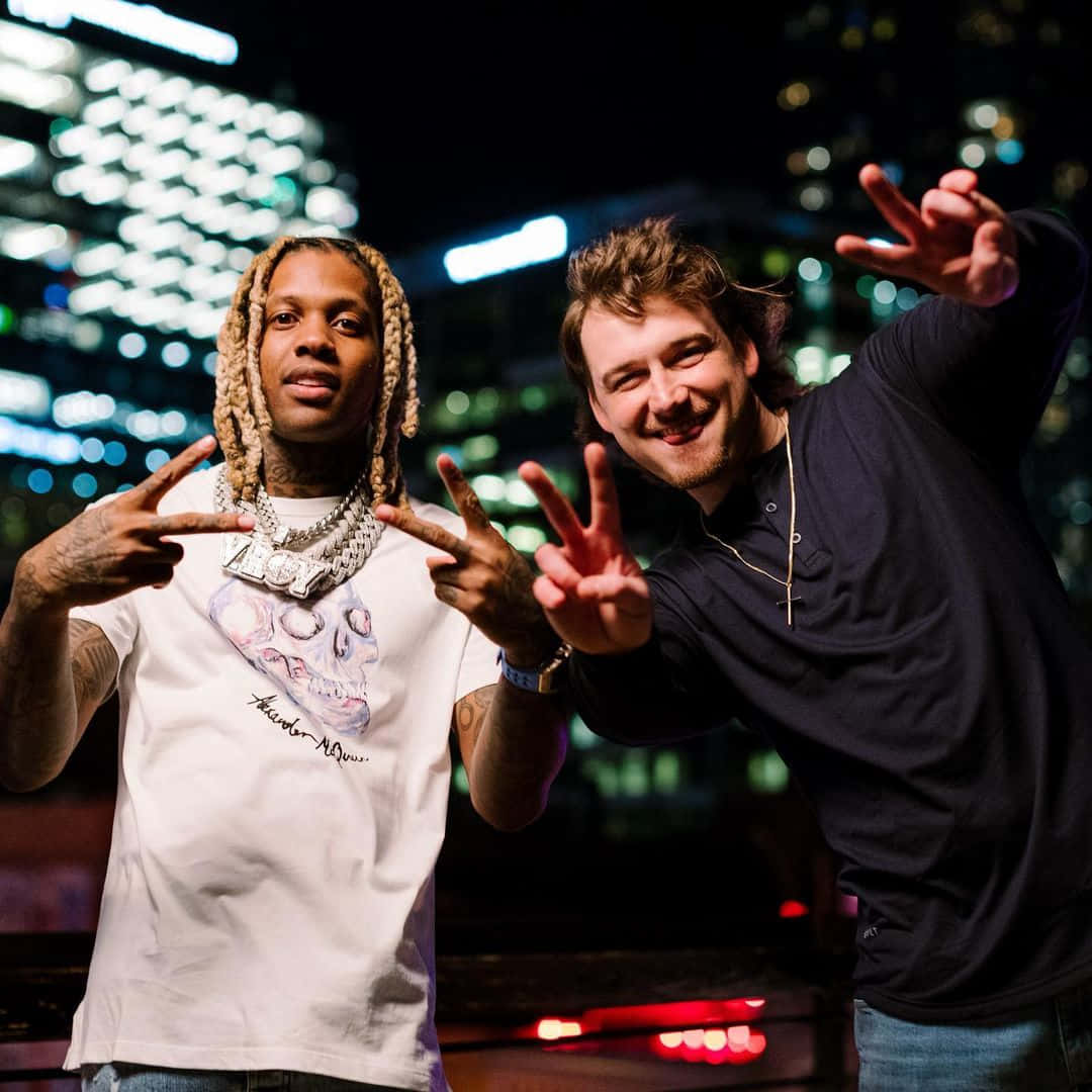 Lil Durk 桌布 壁紙 — 兩名男子在夜間充滿活力的散景燈光城市天際線前擺出和平手勢。