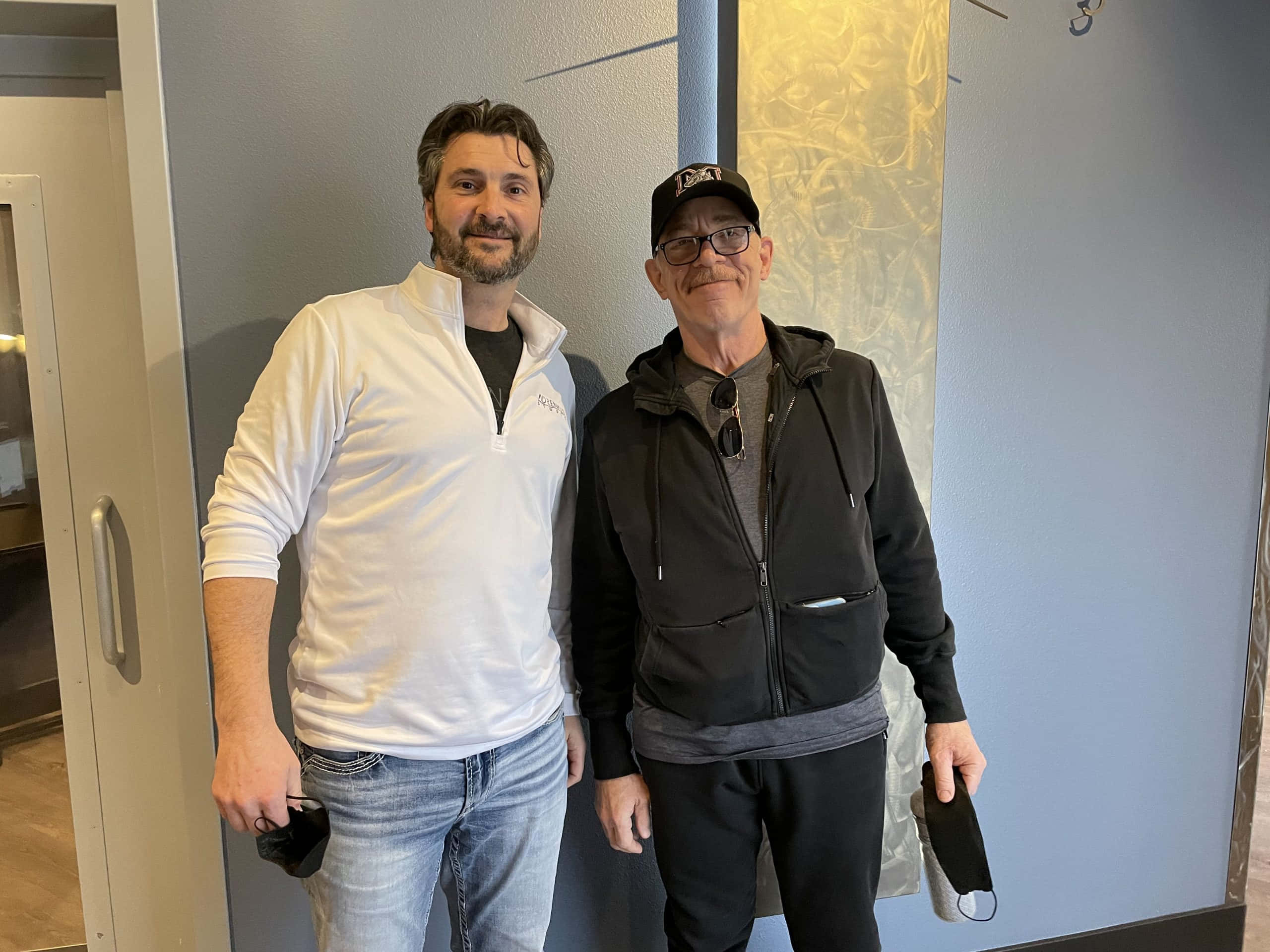 J.k. Simmons 桌布 壁紙 — 兩名男子在室內微笑站立，身後是現代風格的牆壁，帶有金色紋理面板。