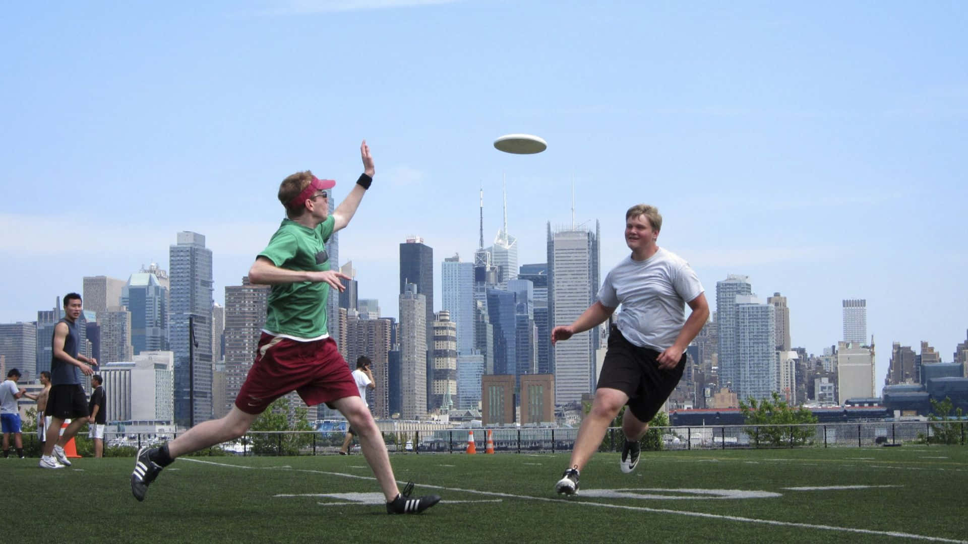 HD Frisbee 桌布 壁紙 — 兩名男子在屋頂場地上跳躍並伸手去接飛行的飛盤，身後是高聳的城市天際線。