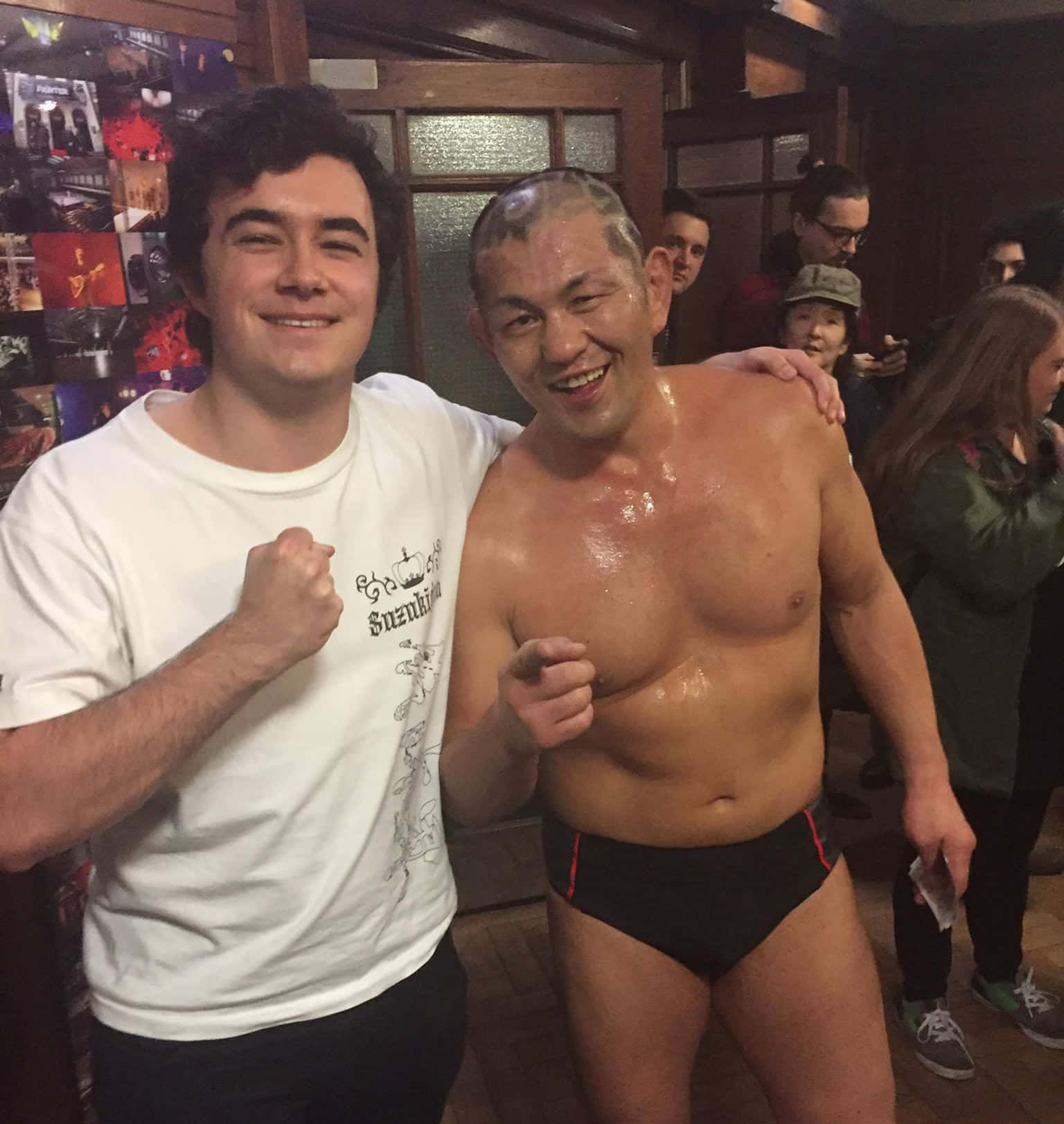 Minoru Suzuki 桌布 壁紙 — 兩名男子在溫暖照明的室內環境中擺出勝利的笑容，一人赤裸上身且汗流浹背，另一人穿著白色T恤，周圍有旁觀者。