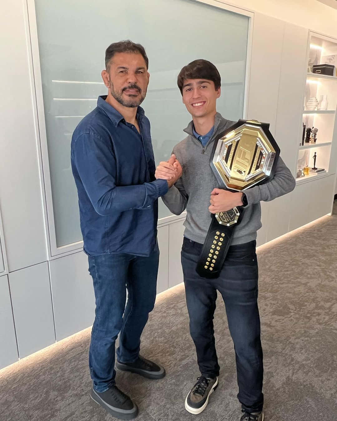 UFC 桌布 壁紙 — 兩名男子在現代辦公室中，一人手持UFC冠軍腰帶，與微笑的年輕男子握手。