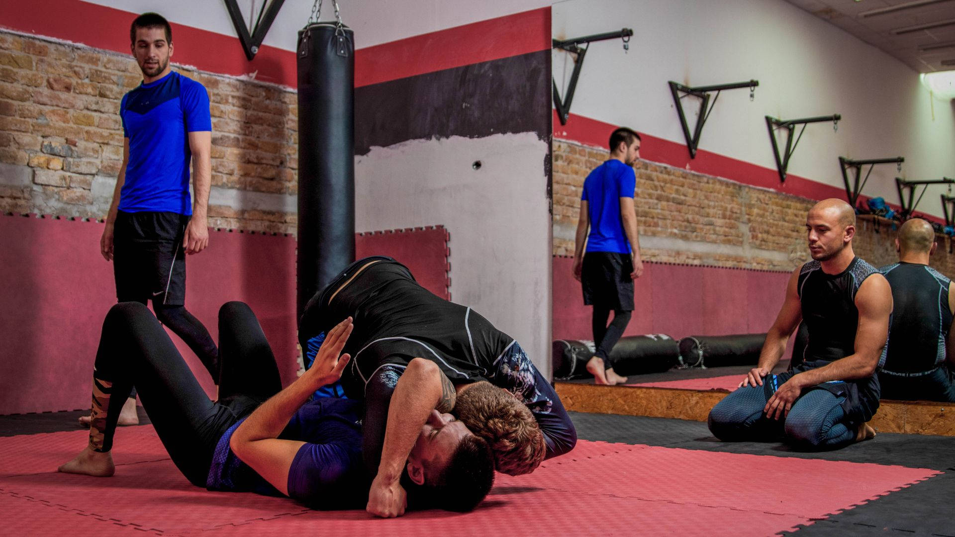 martial arts 桌布 壁紙 — 兩名男子在磚牆健身房的紅色墊子上激烈纏鬥，其他人則在旁觀看或附近訓練。