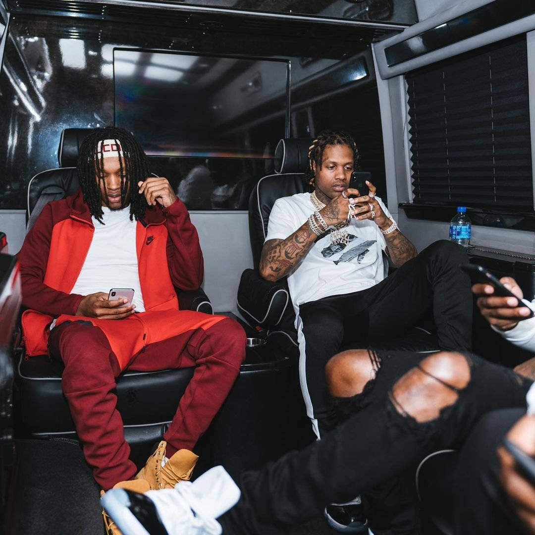 King Von And Lil Durk 桌布 壁紙 — 兩名男子在豪華廂型車內休息，一人身穿紅色運動服，兩人都專注於手機，刺青可見，皮革座椅，環境照明。