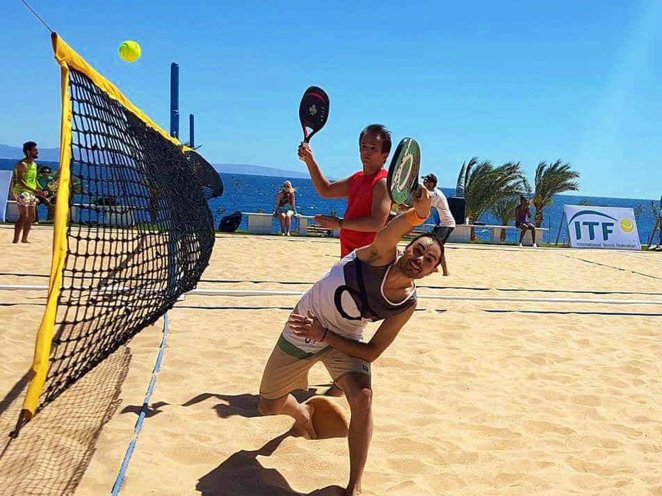 Beach Tennis 桌布 壁紙 — 兩名男子在陽光明媚的日子裡，以海洋和棕櫚樹為背景，激烈地進行沙灘網球比賽。