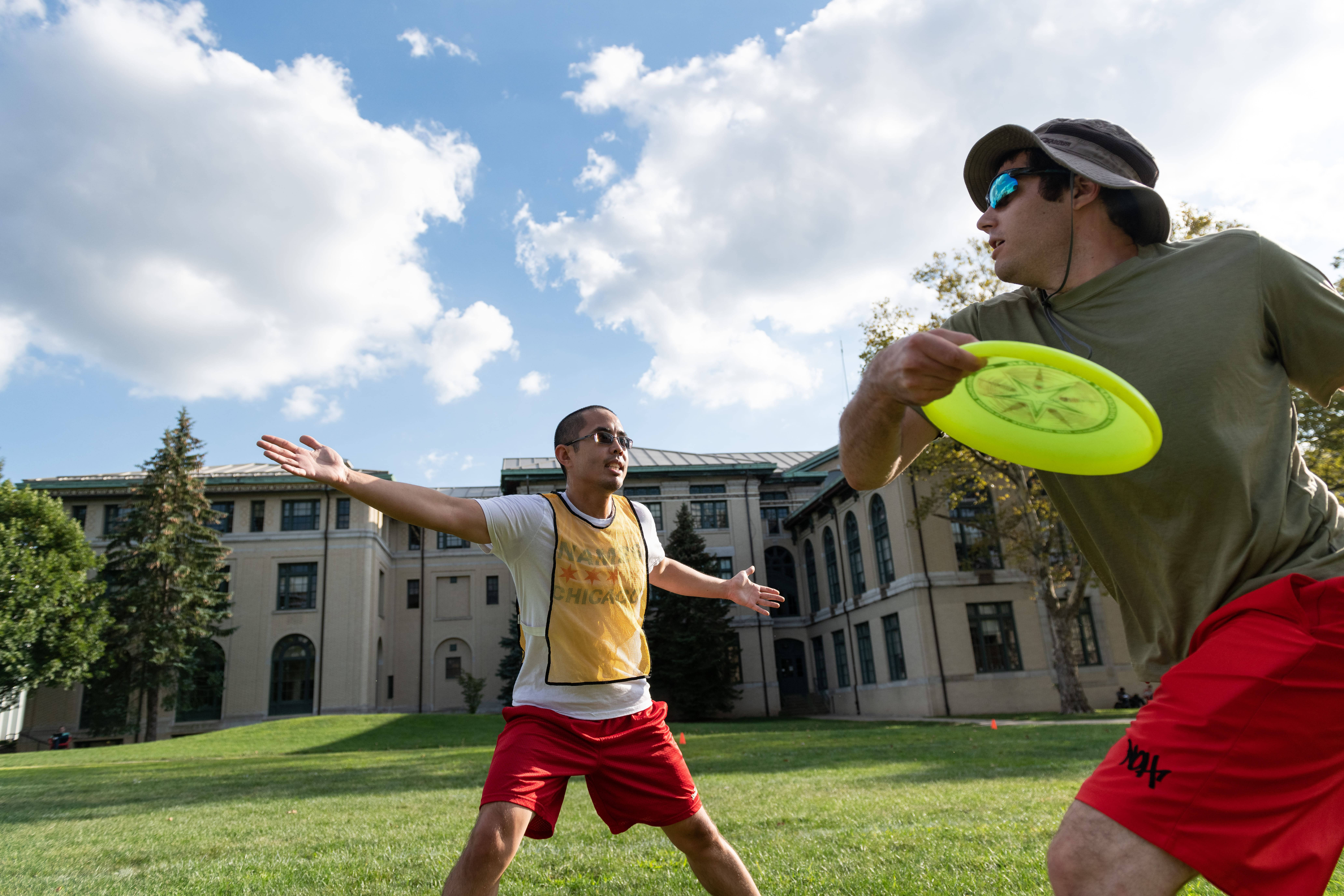 Frisbee 桌布 壁紙 — 兩名男子在陽光明媚的校園草坪上玩飛盤，身後是一座宏偉的建築，頭頂是明亮的藍天。