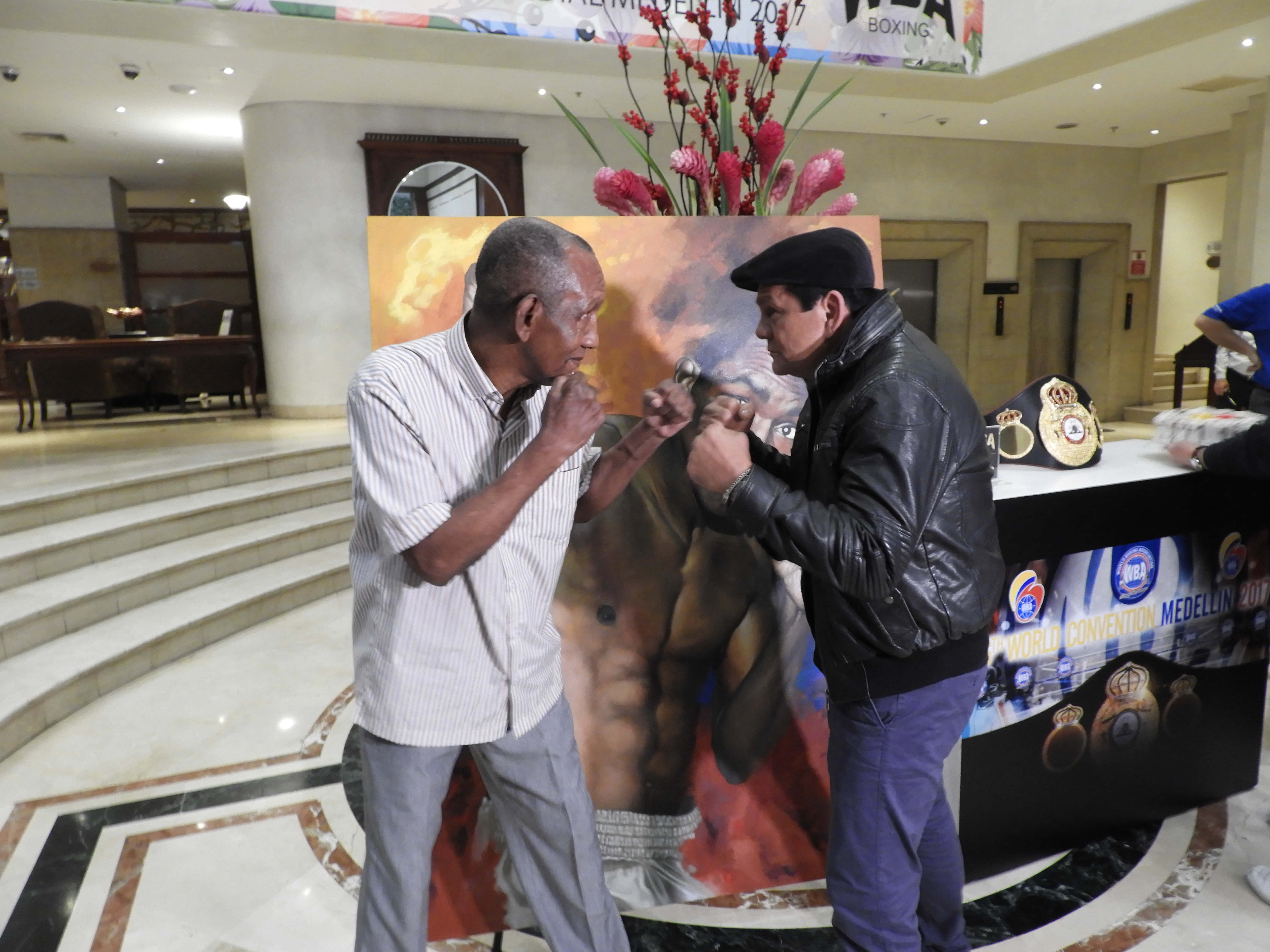 Boxing 桌布 壁紙 — 兩名男子在飯店大廳戴著手套模仿拳擊比賽，背景是一張肌肉發達的拳擊手海報。