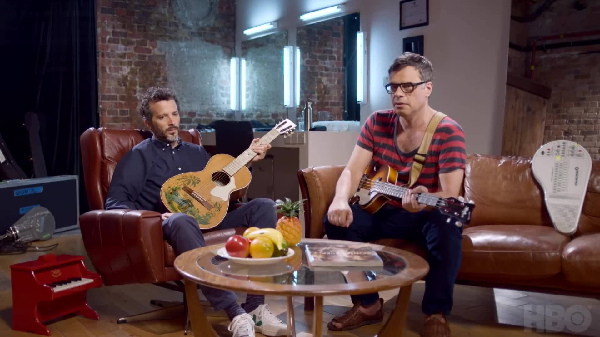 Flight Of The Conchords 桌布 壁紙 — 兩名男子坐在一個質樸的磚牆房間裡，圍著一張放有水果的咖啡桌彈奏原聲吉他和烏克麗麗。