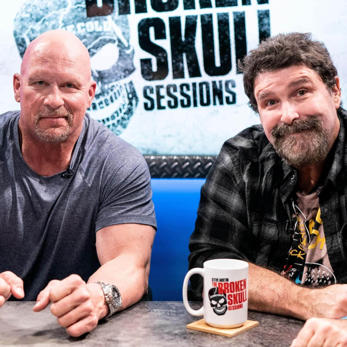 WWE 桌布 壁紙 — 兩名男子坐在桌子旁，桌上有一個杯子，背景寫著「Broken Skull Sessions」，並帶有骷髏標誌。