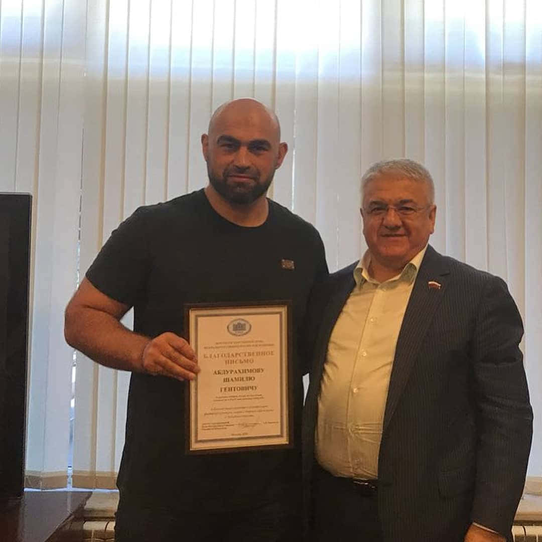 MMA 桌布 壁紙 — 兩名男子室內站立，一人手持帶有西里爾文字的裱框證書，背景為垂直百葉窗。