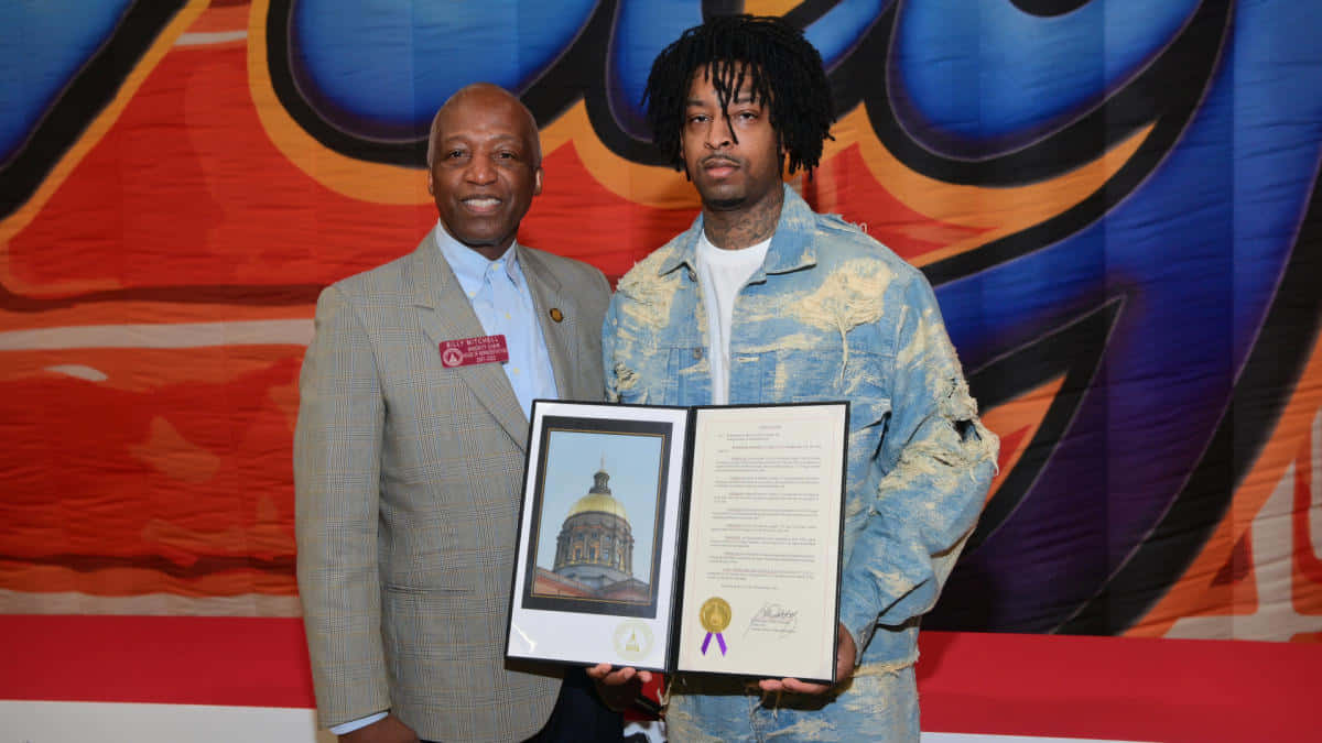 21 Savage 桌布 壁紙 — 兩名男子微笑著手持裱框證書和肖像，背景是充滿活力的塗鴉風格。