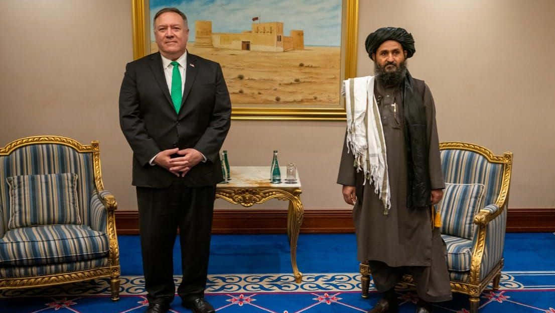 Taliban 桌布 壁紙 — 兩名男子正式地站在一個華麗的房間裡，房間裡有藍色圖案地毯和條紋椅子，一人身穿西裝打綠色領帶，另一人身穿傳統長袍和頭巾。