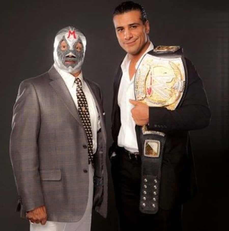 WWE 桌布 壁紙 — 兩名男子正式擺姿勢，一人身穿灰色西裝，戴著帶有紅色M標誌的白色面具，另一人身穿黑色西裝，手持冠軍腰帶，背景為深色攝影棚。