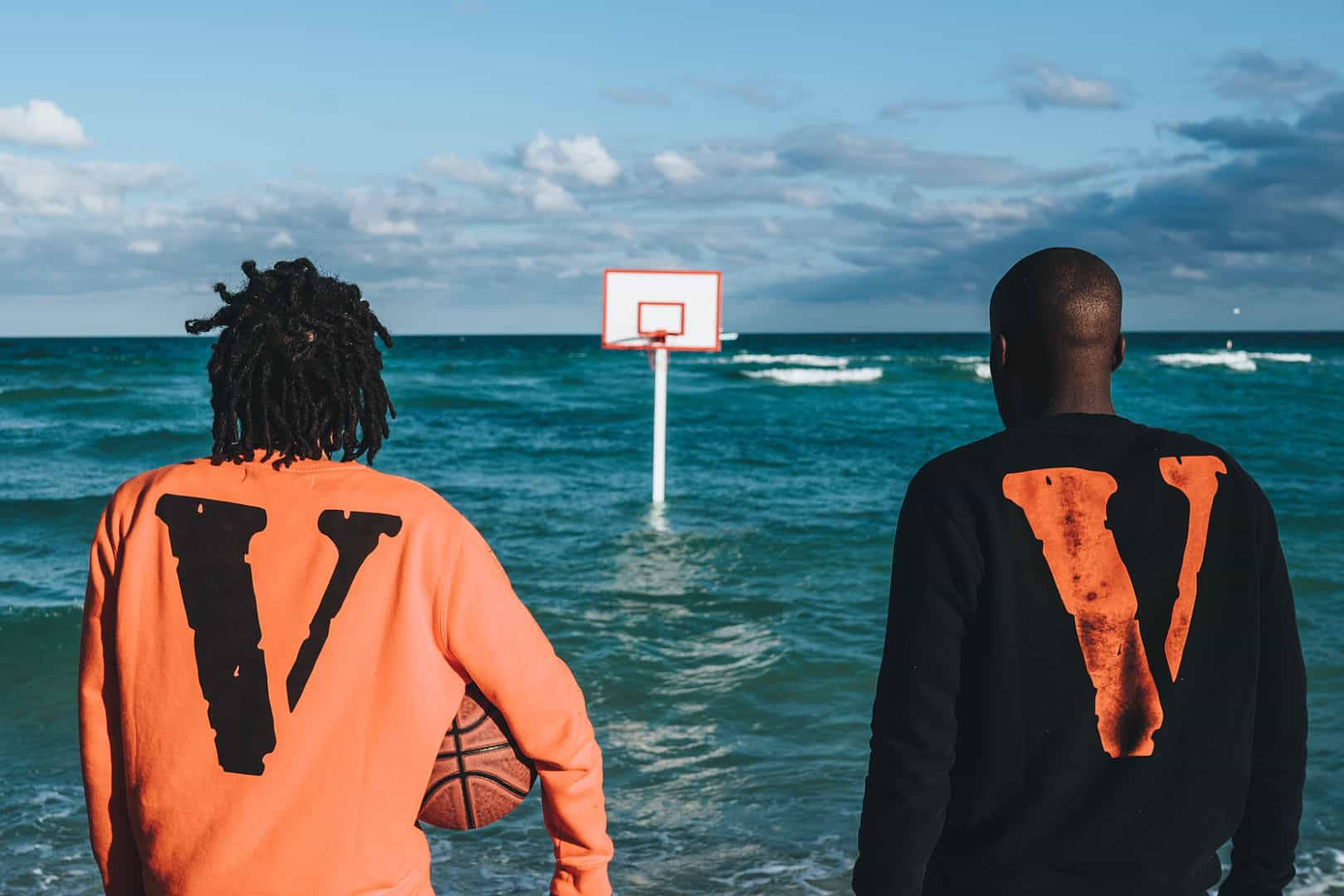 Vlone 桌布 壁紙 — 兩名男子穿著相配的「V」連帽衫，站在齊腰深的海水中，面對著一個部分多雲的天空下浸沒的籃球框。