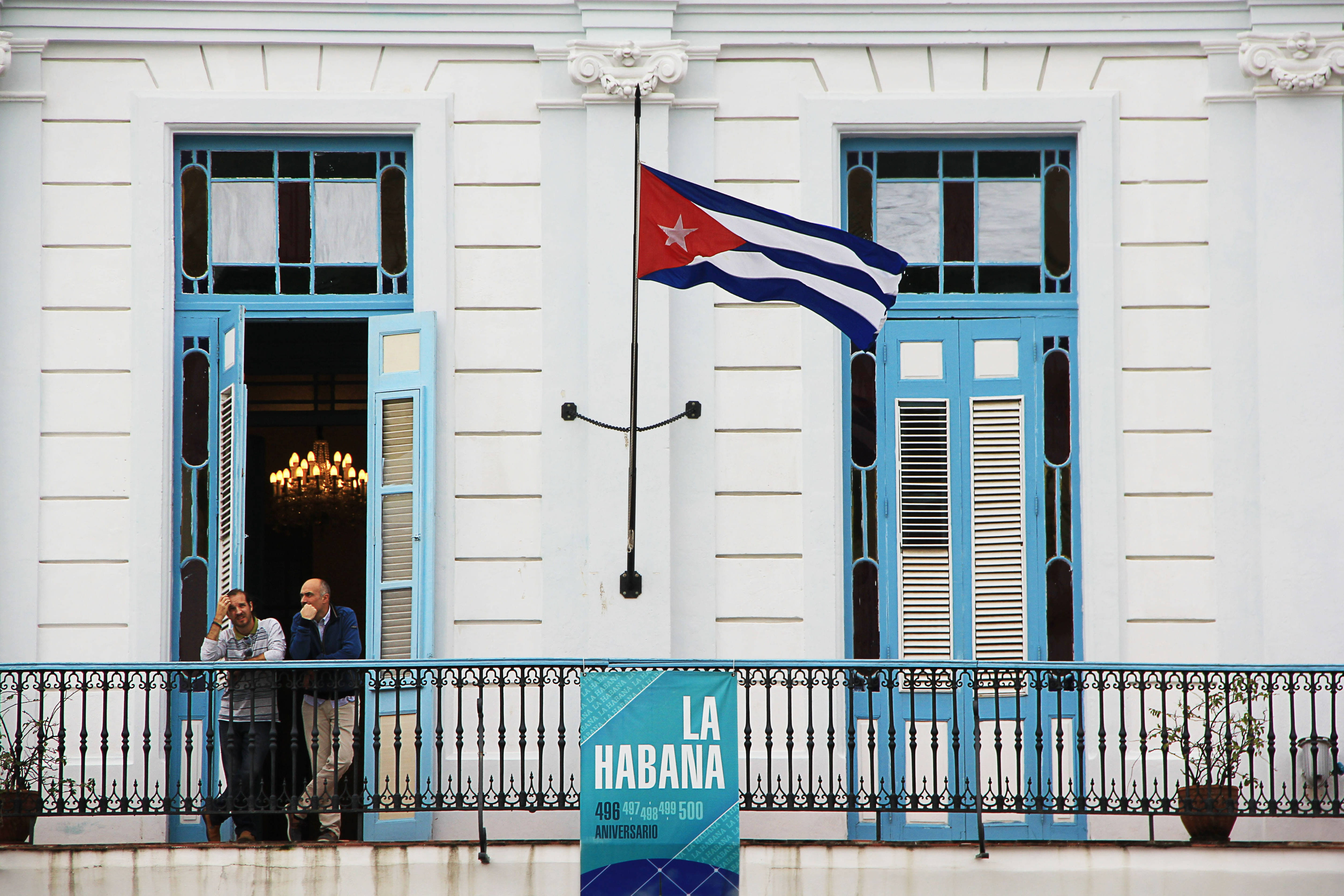 Cuban 桌布 壁紙 — 兩名男子站在一棟帶有藍色百葉窗的白色殖民風格建築的陽台上，一面古巴國旗在他們上方飄揚。