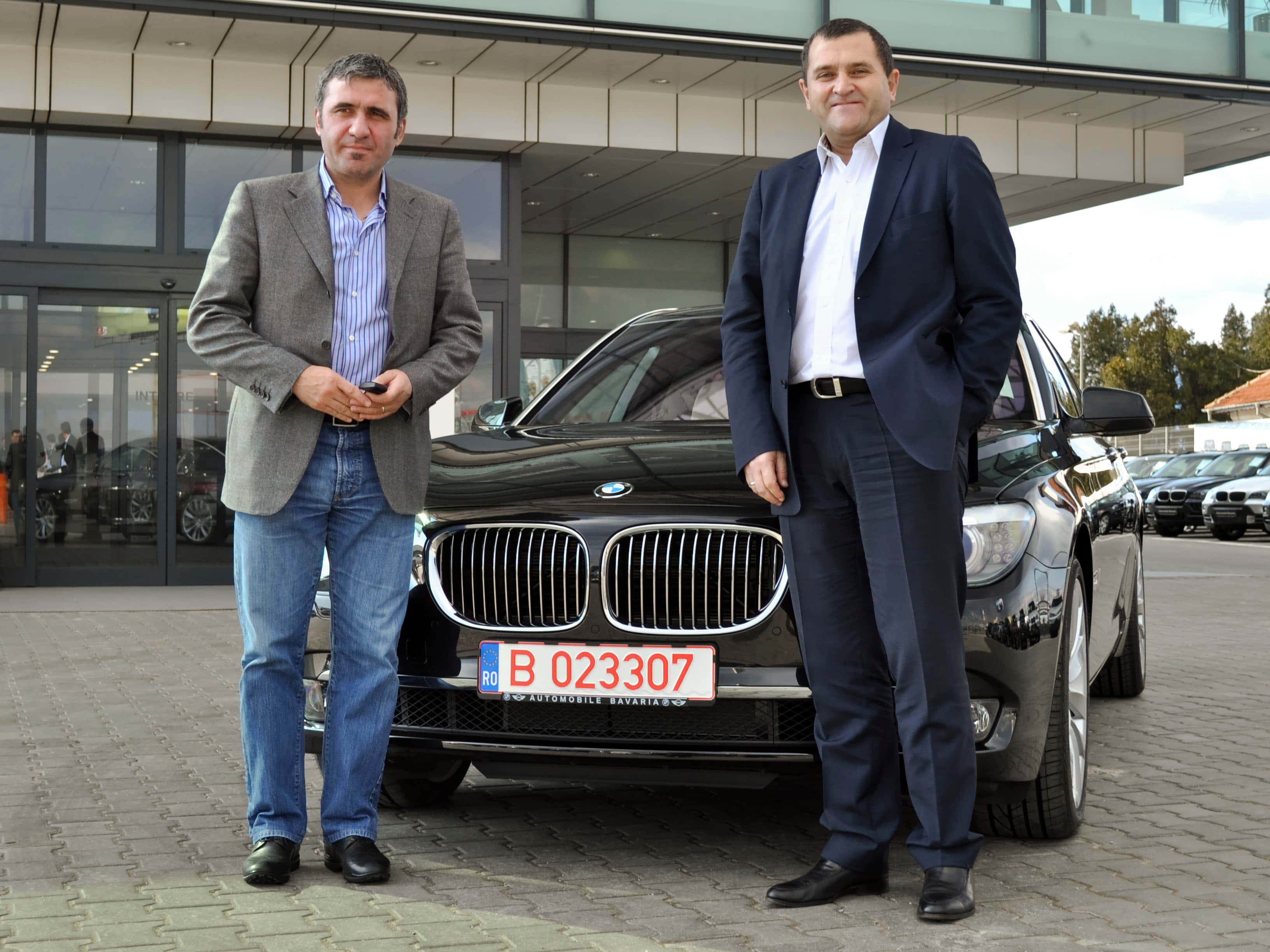 BMW 動圖 桌布 壁紙 — 兩名男子自信地站在一輛時尚的黑色BMW SUV旁邊，地點是現代汽車經銷商，擁有玻璃外牆和鋪砌的庭院。