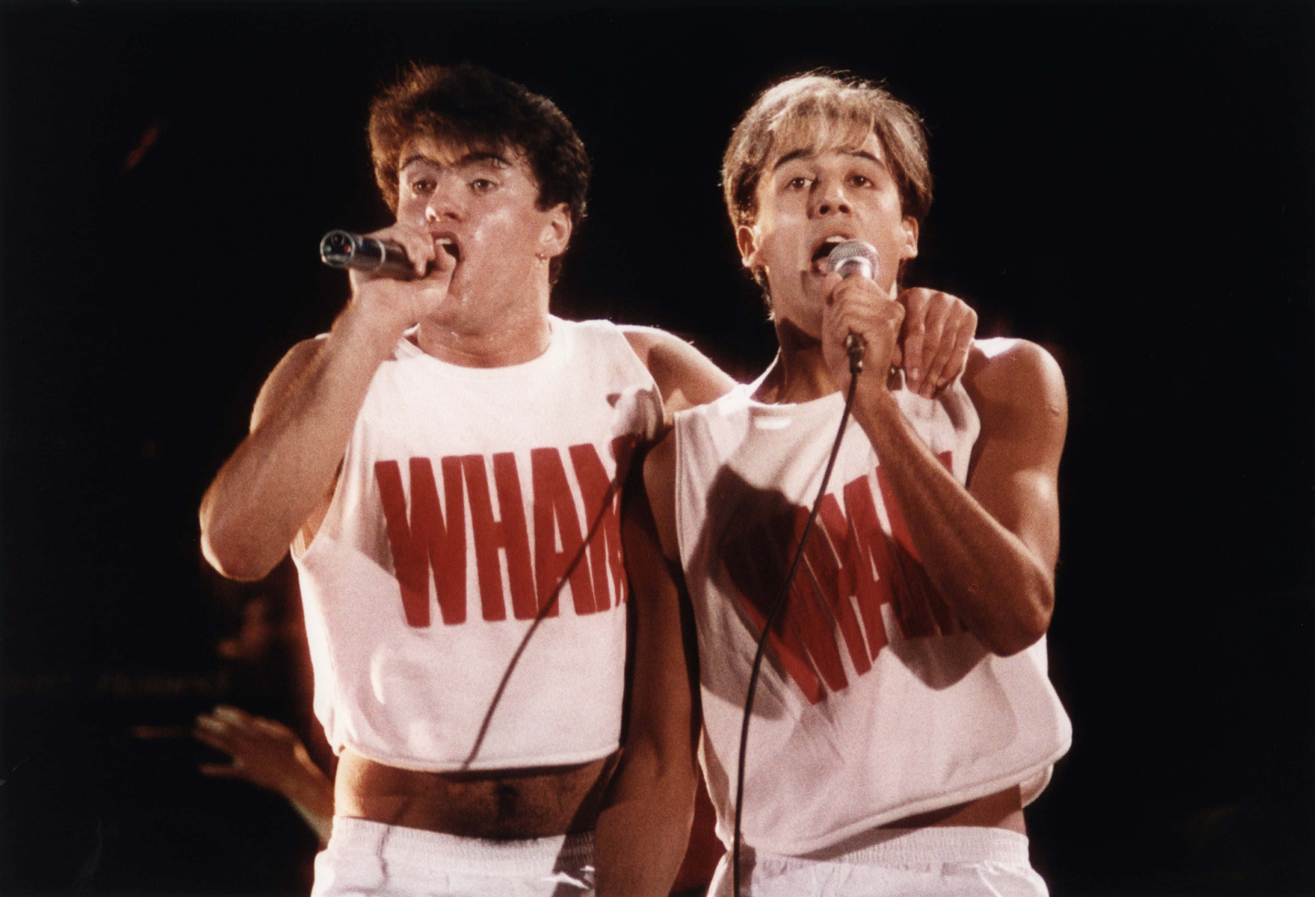 band 桌布 壁紙 — 兩名男歌手身穿印有紅色「WHAM」字樣的白色背心，在明亮的舞台燈光下手持麥克風充滿活力地表演。