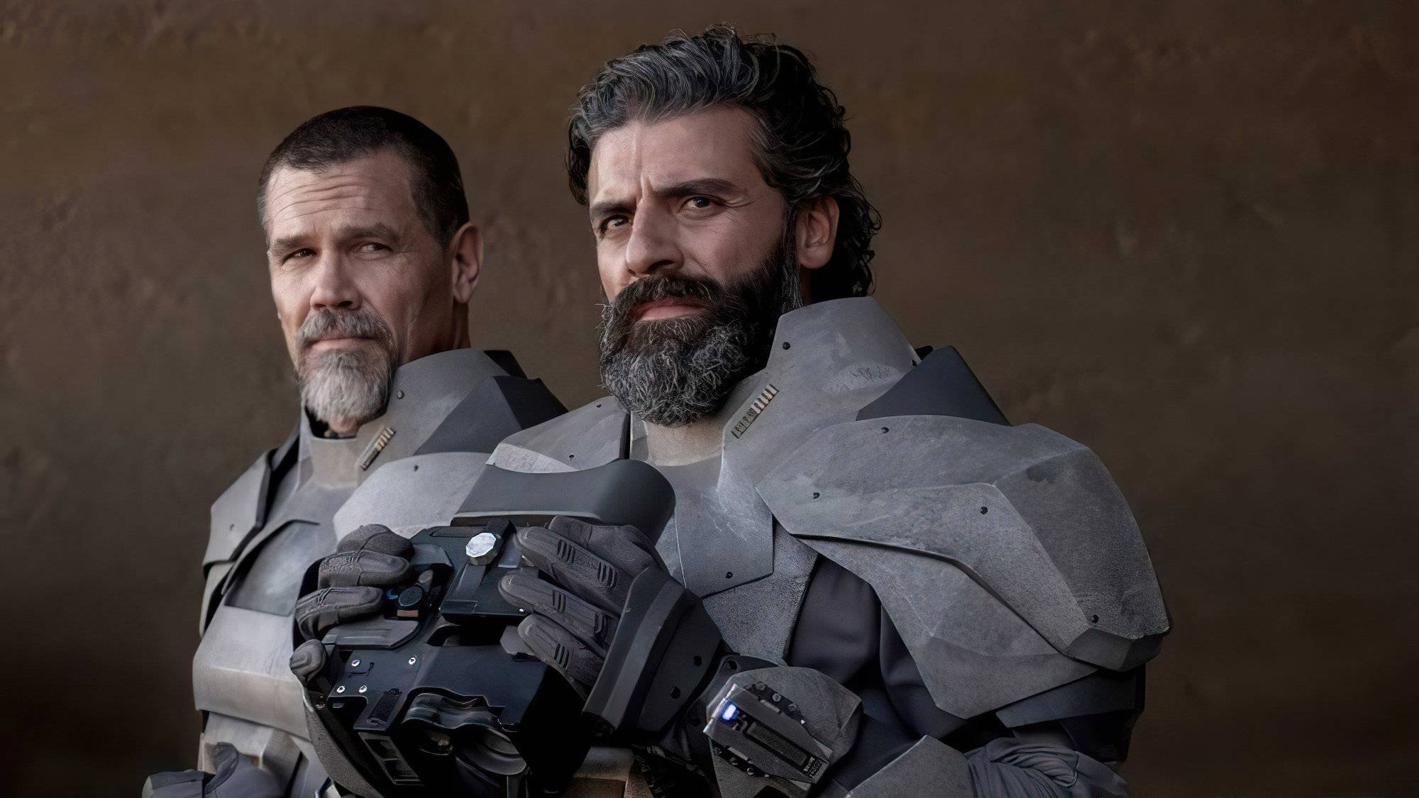Dune Movie 桌布 壁紙 — 兩名留鬍鬚的男子身穿未來感灰色盔甲，莊嚴地站在有紋理的棕色背景前，其中一人手持高科技武器。
