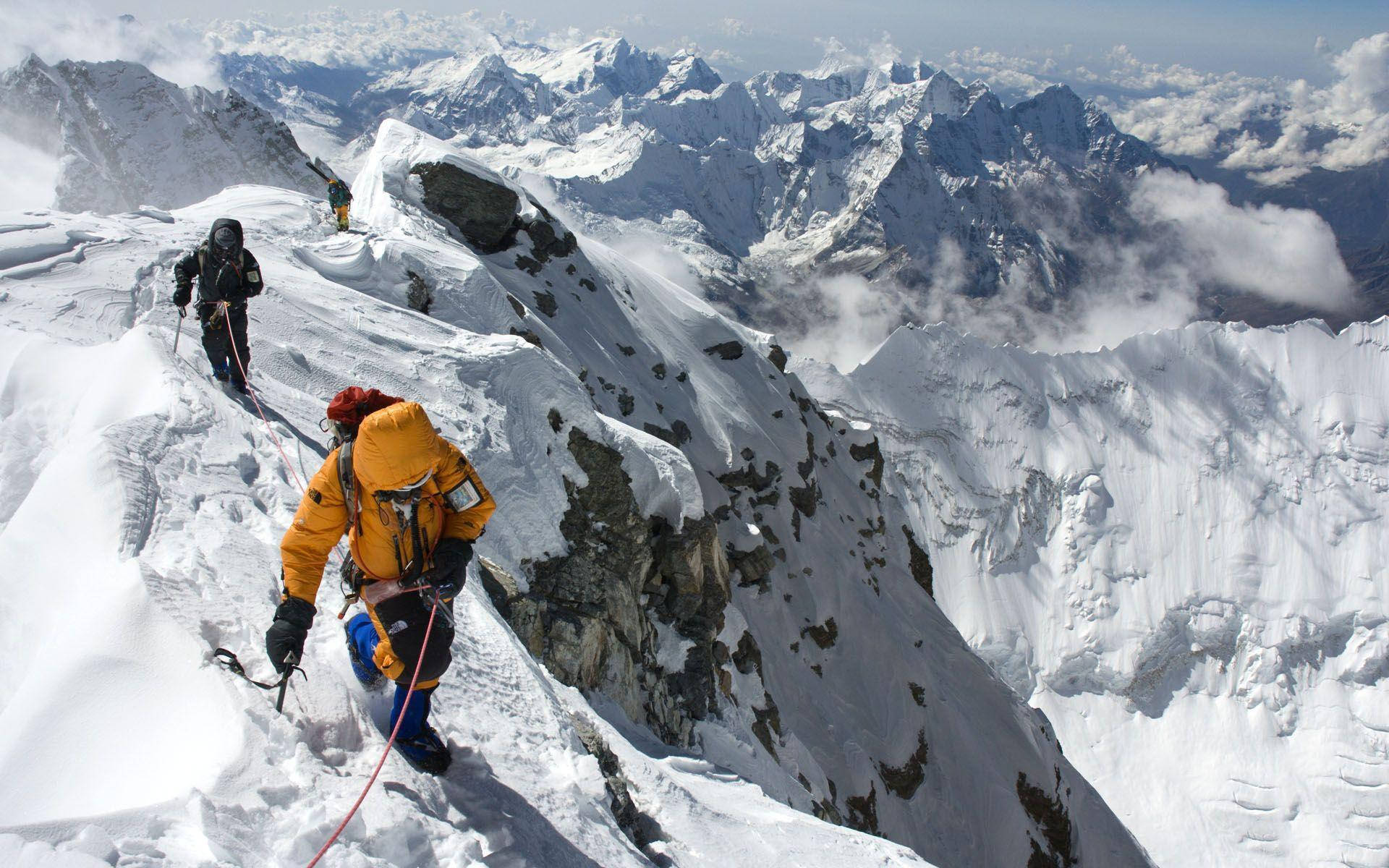山景 桌布 壁紙 — 兩名登山者攀登一條危險的積雪山脊，下方是廣闊的雪峰山脈和雲層。