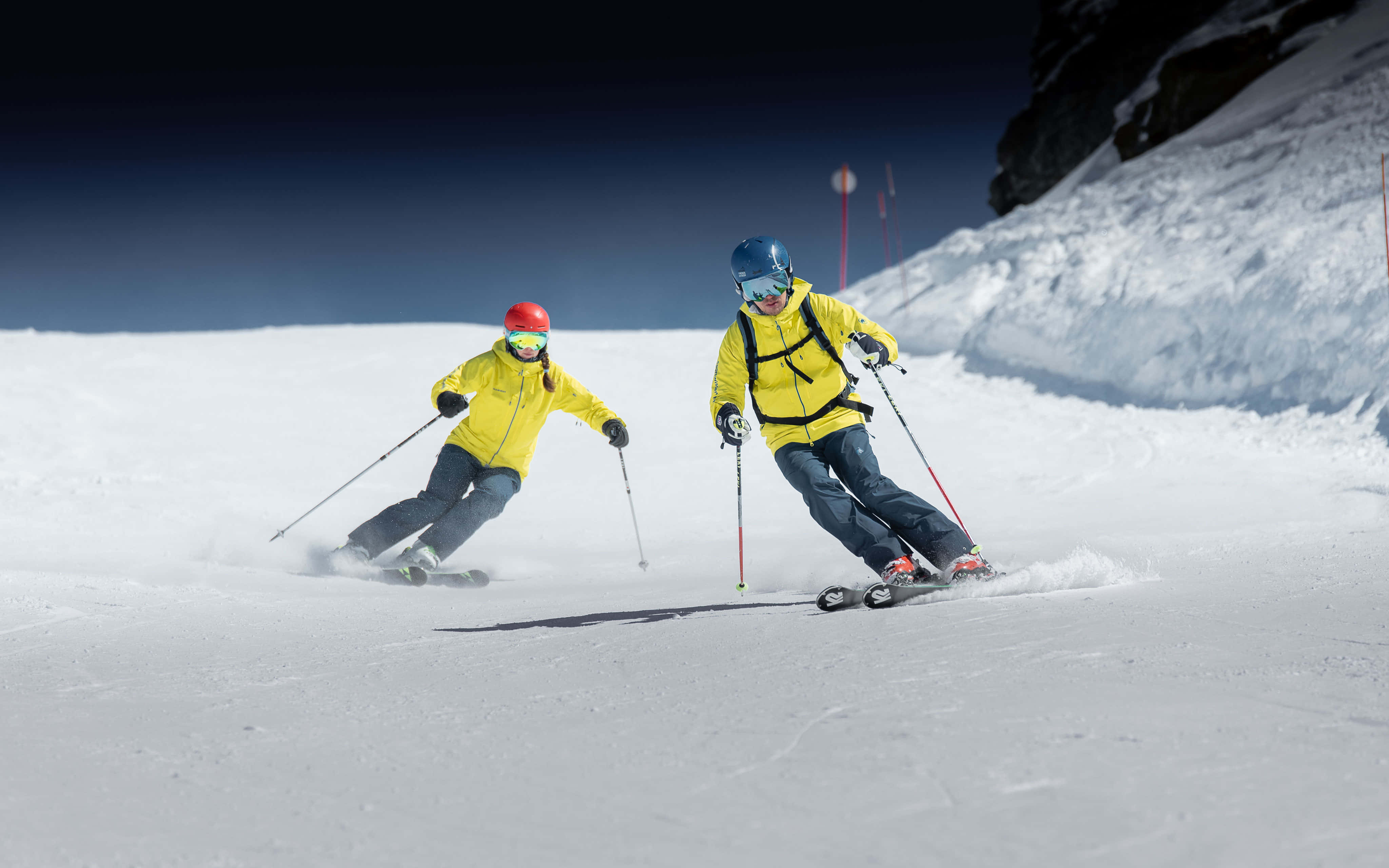 ski 桌布 壁紙 — 兩名穿著亮黃色外套的滑雪者，在晴朗的藍天下沿著雪坡滑行。