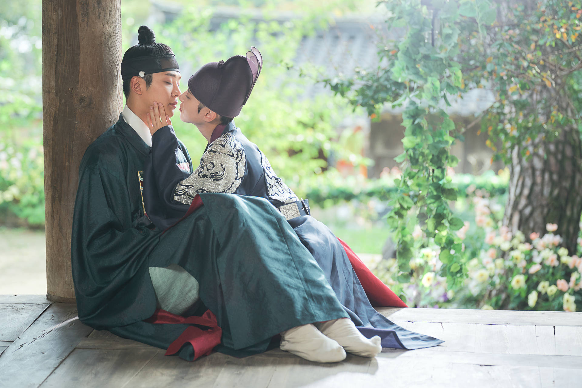 kdrama 桌布 壁紙 — 兩名穿著傳統韓國韓服的男士在木製門廊上分享親密時刻，周圍環繞著茂密的綠植和盛開的花朵。