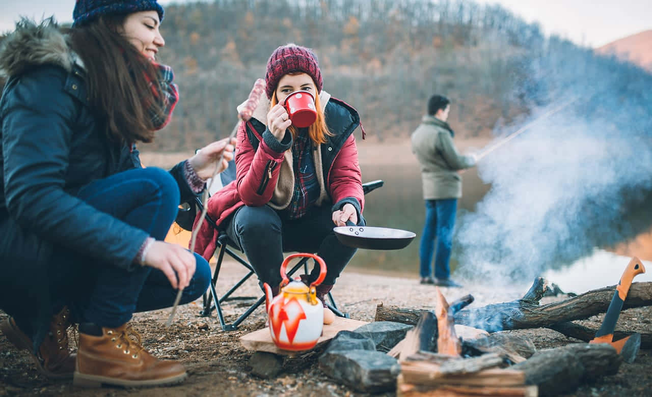 Fall Camping 桌布 壁紙 — 兩名穿著冬季裝備的女性在營火旁享受熱飲，煙霧升騰，一名男子在附近的森林環境中烹飪。