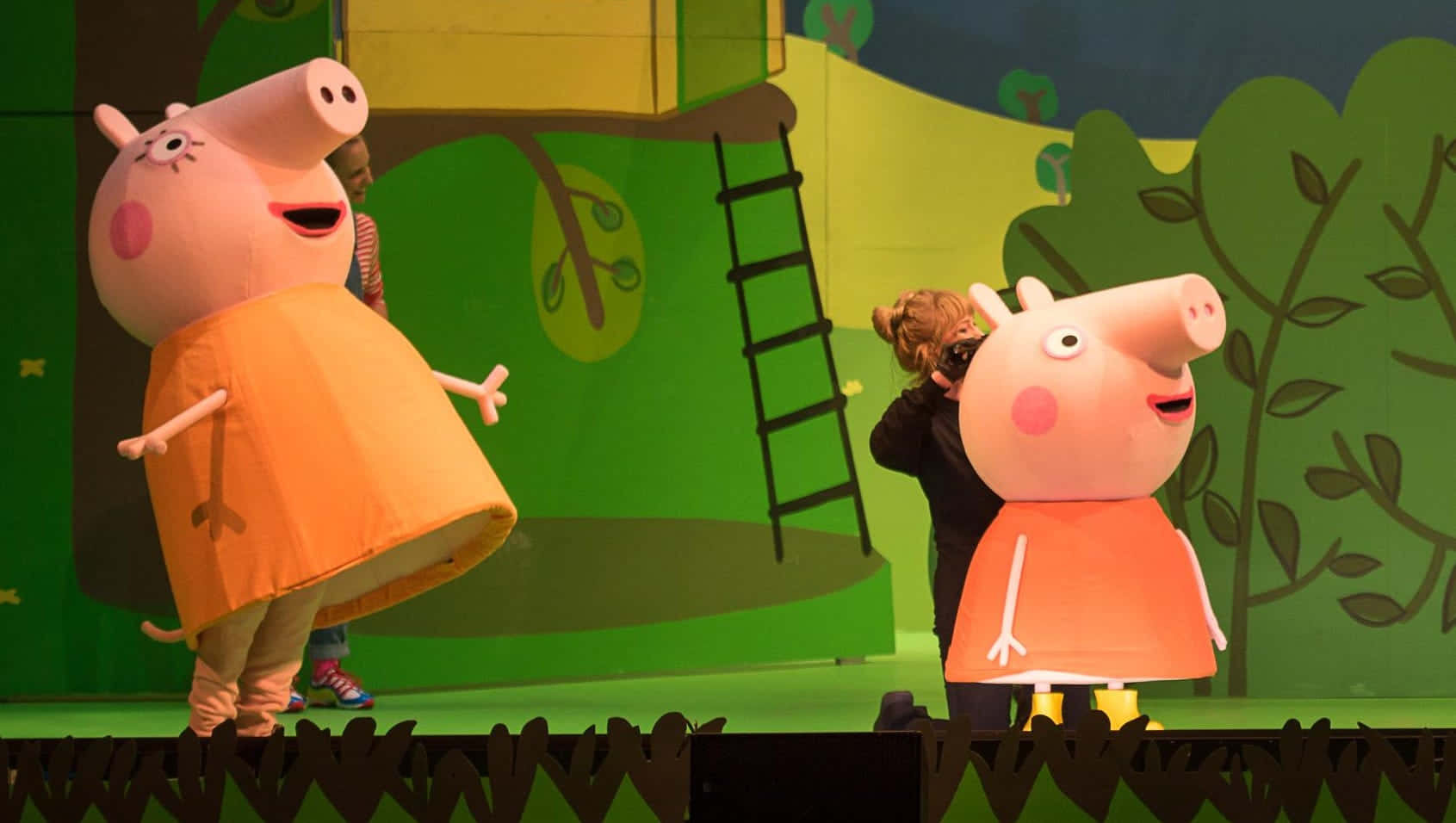 Pedro Pony 桌布 壁紙 — 兩名穿著大型Peppa Pig服裝的表演者站在一個有樹木和梯子繪畫的綠色背景舞台上。