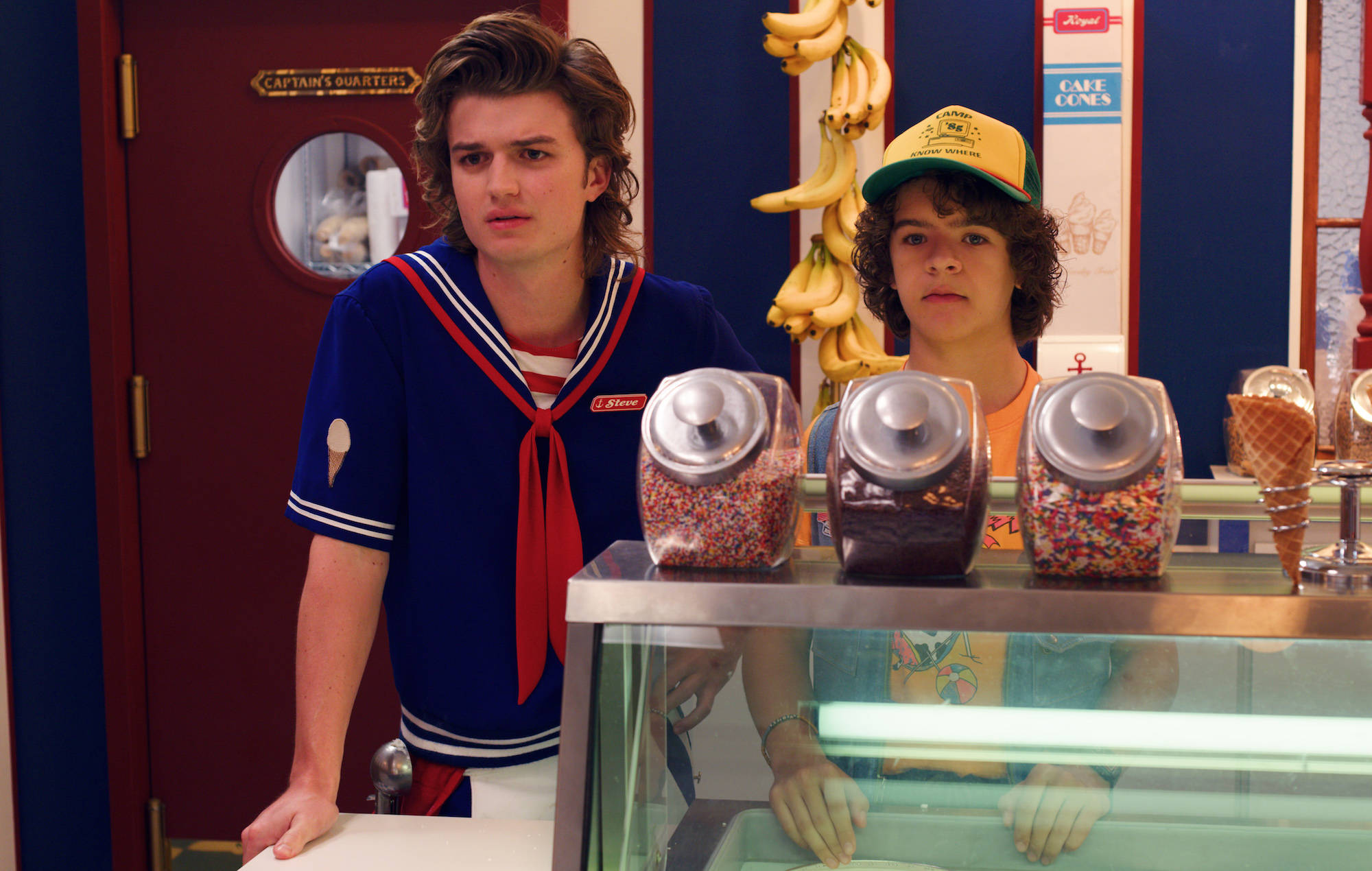 Dustin Stranger Things 桌布 壁紙 — 兩名穿著復古服裝的年輕男子站在一個冰淇淋櫃檯後面，櫃檯上方掛著彩色糖粒和香蕉。