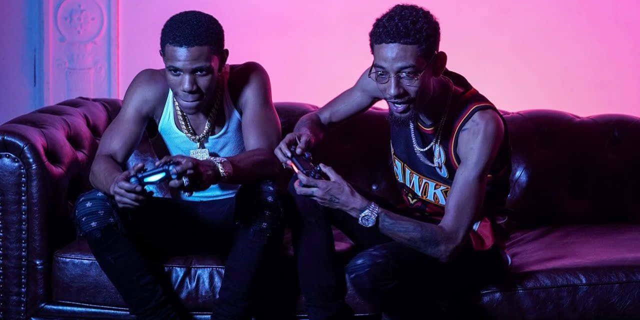 A Boogie Wit Da Hoodie 桌布 壁紙 — 兩名穿著時尚街頭服的男士在充滿活力的粉紅和藍色霓虹燈光下，在皮革沙發上激烈地玩電子遊戲。