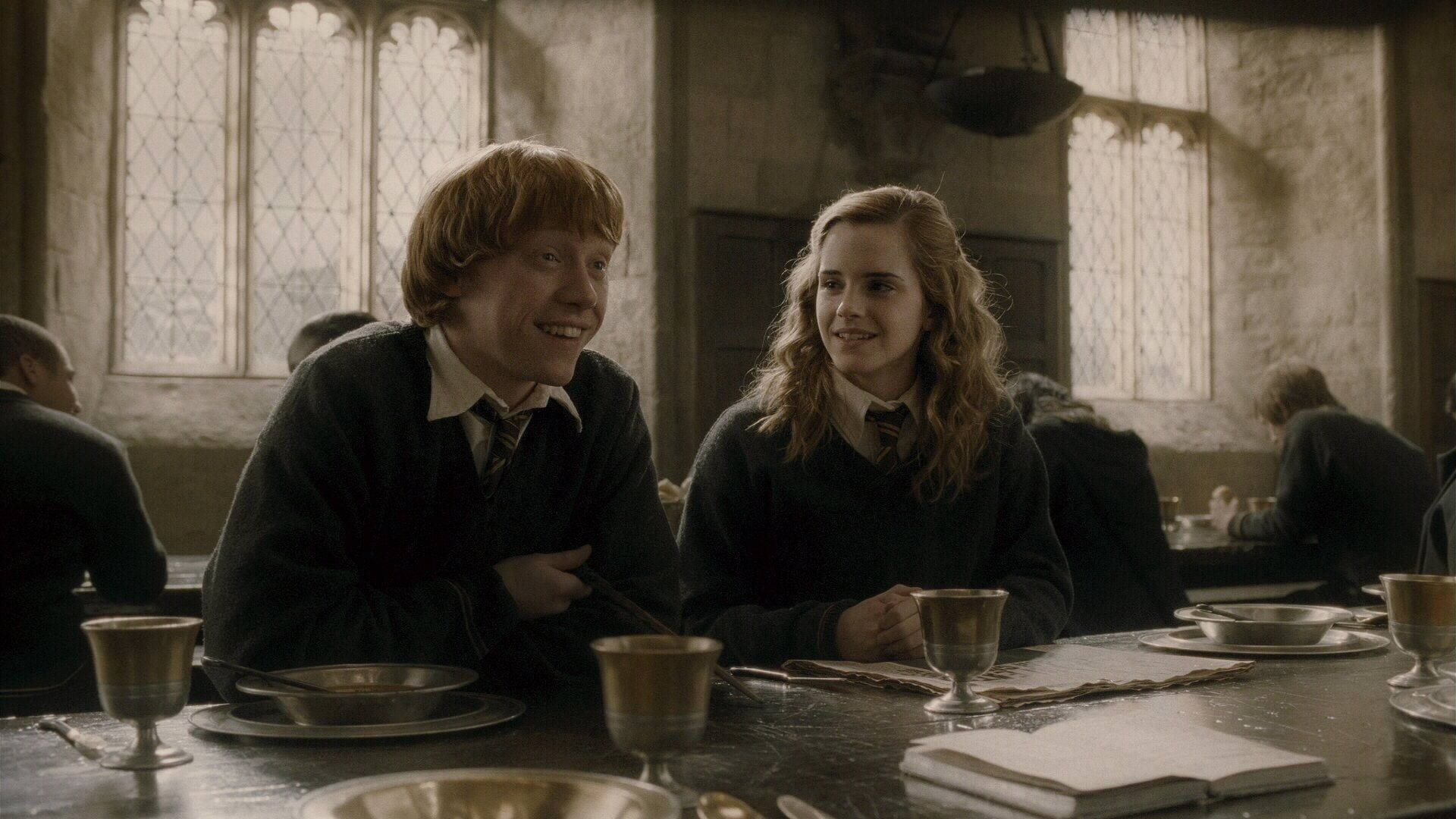 Ron Weasley 桌布 壁紙 — 兩名穿著校服的學生在一個有石牆和彩繪玻璃窗的大廳裡，在一張長餐桌上分享溫暖的笑容。