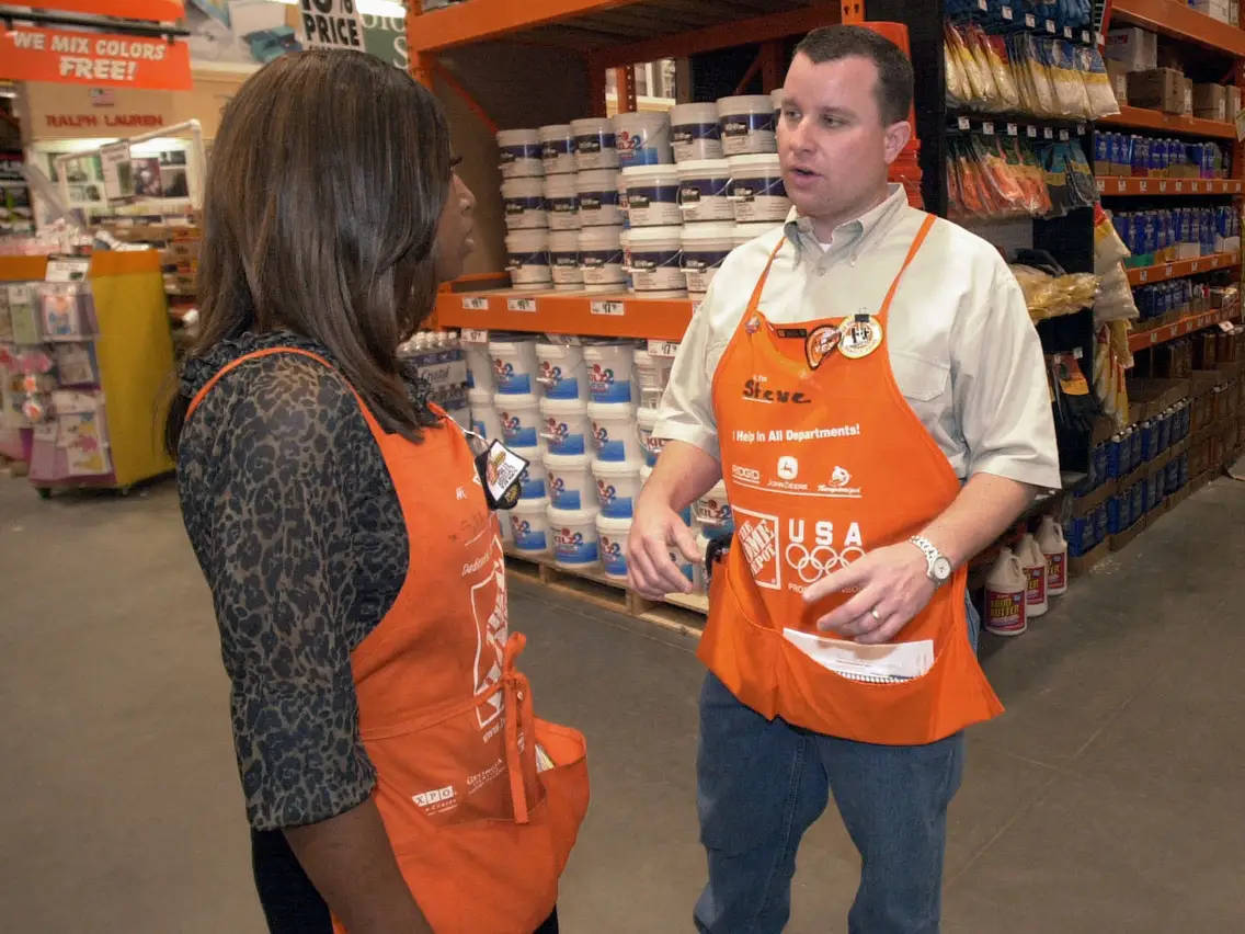 Home Depot 桌布 壁紙 — 兩名穿著橙色Home Depot圍裙的員工在光線充足的五金店通道上交談，通道上擺滿了油漆桶。