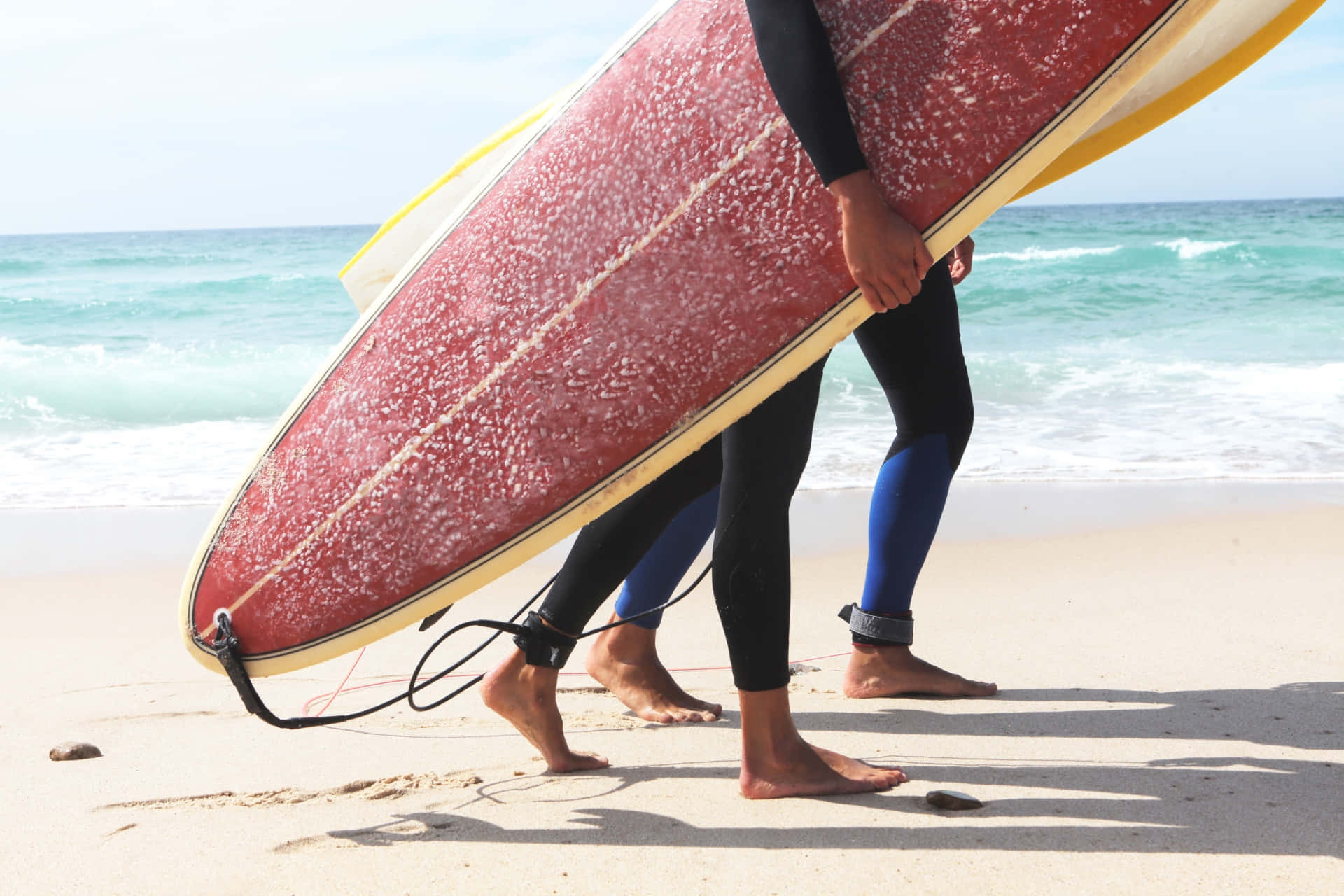 Beach Water Sports 桌布 壁紙 — 兩名穿著潛水衣的衝浪者沿著陽光明媚的沙灘搬運一塊風化的紅色衝浪板，身後有碧綠色的海浪拍打。